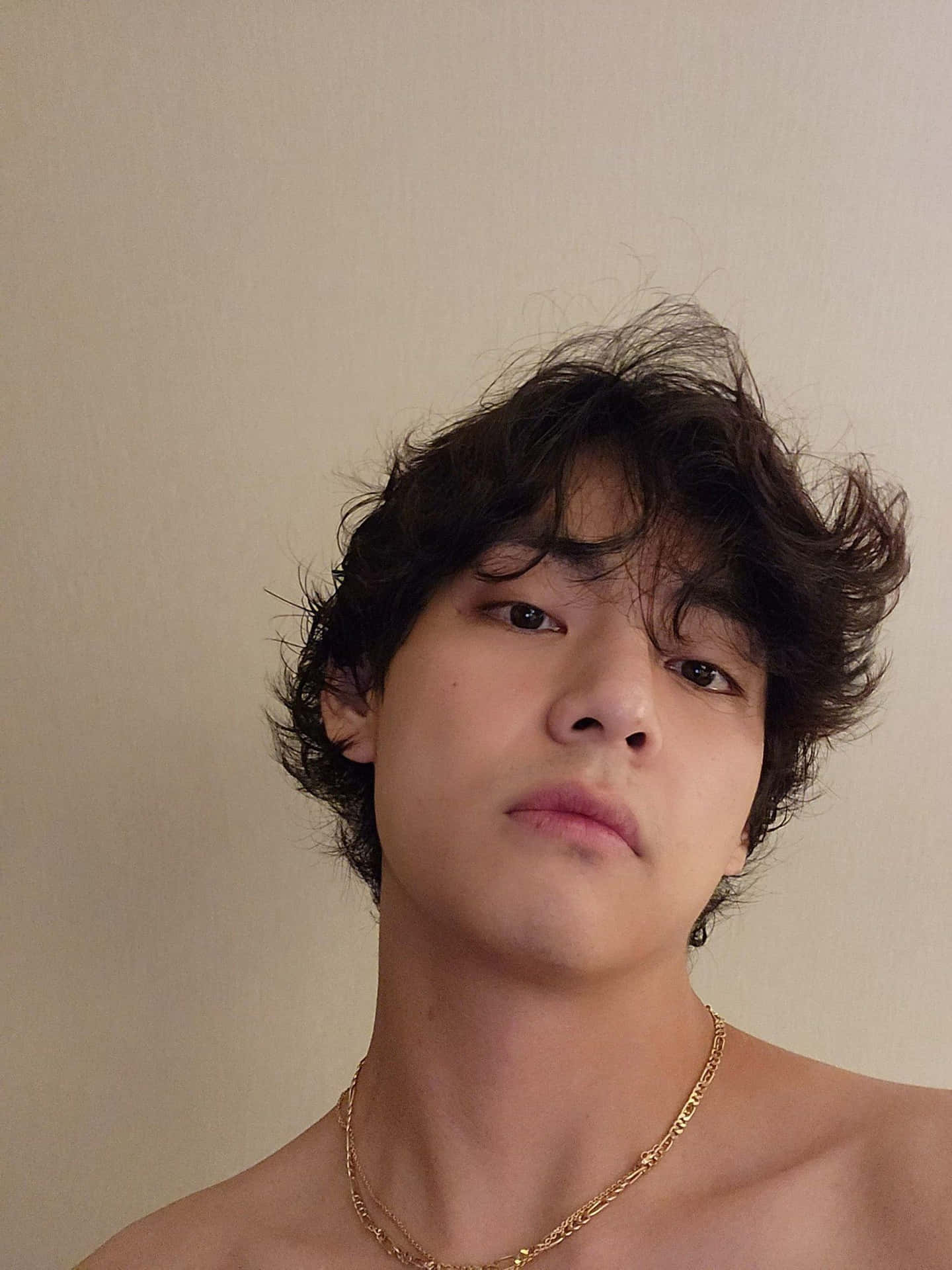  壁紙 — 一個年輕男子，一頭凌亂的深色頭髮，戴著金鍊，在純米色牆壁前直視鏡頭。