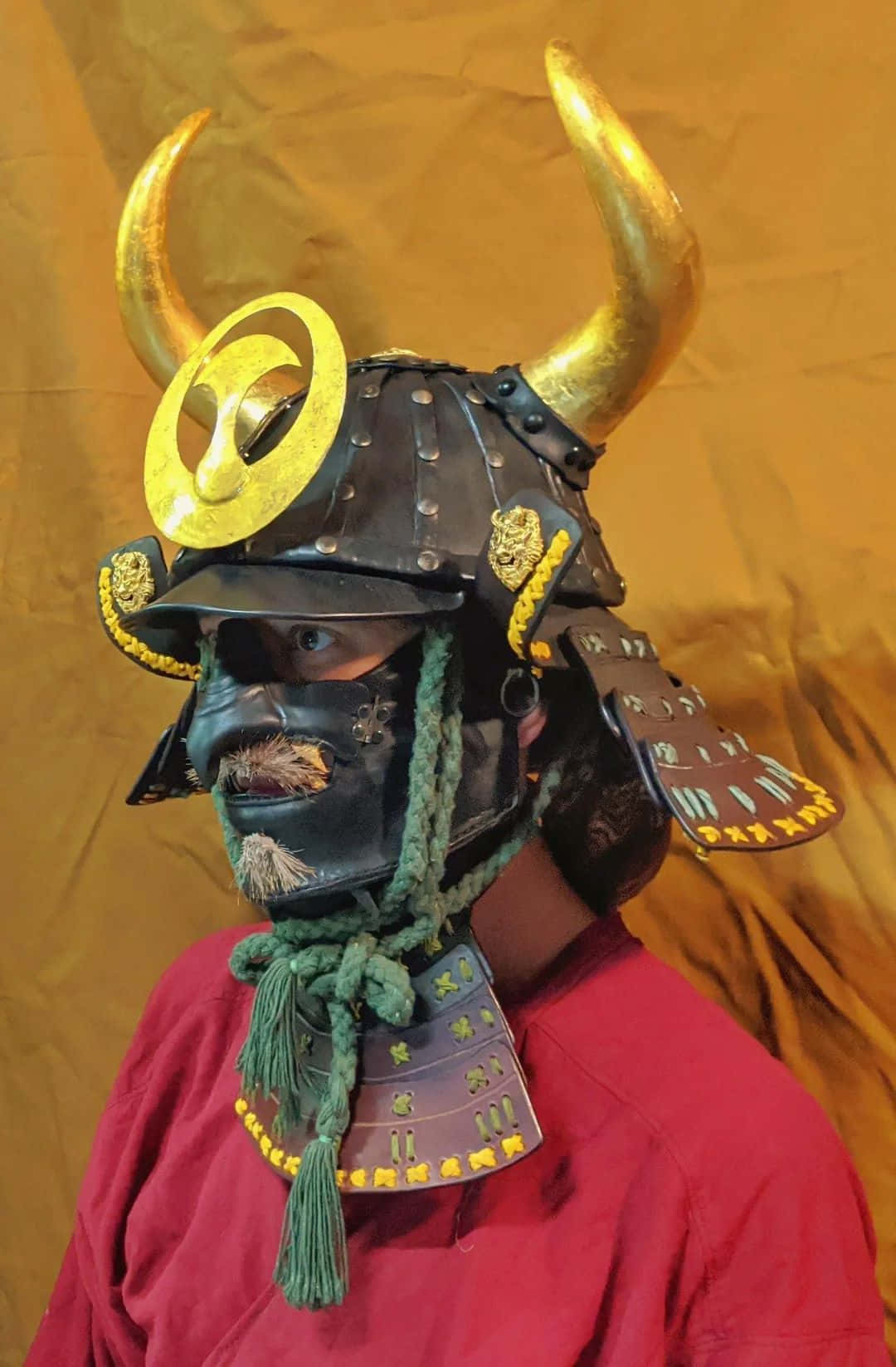  壁紙 — 一個人戴著裝飾華麗的武士頭盔，帶有金色角和綠色繩索，背景是溫暖的橙色。
