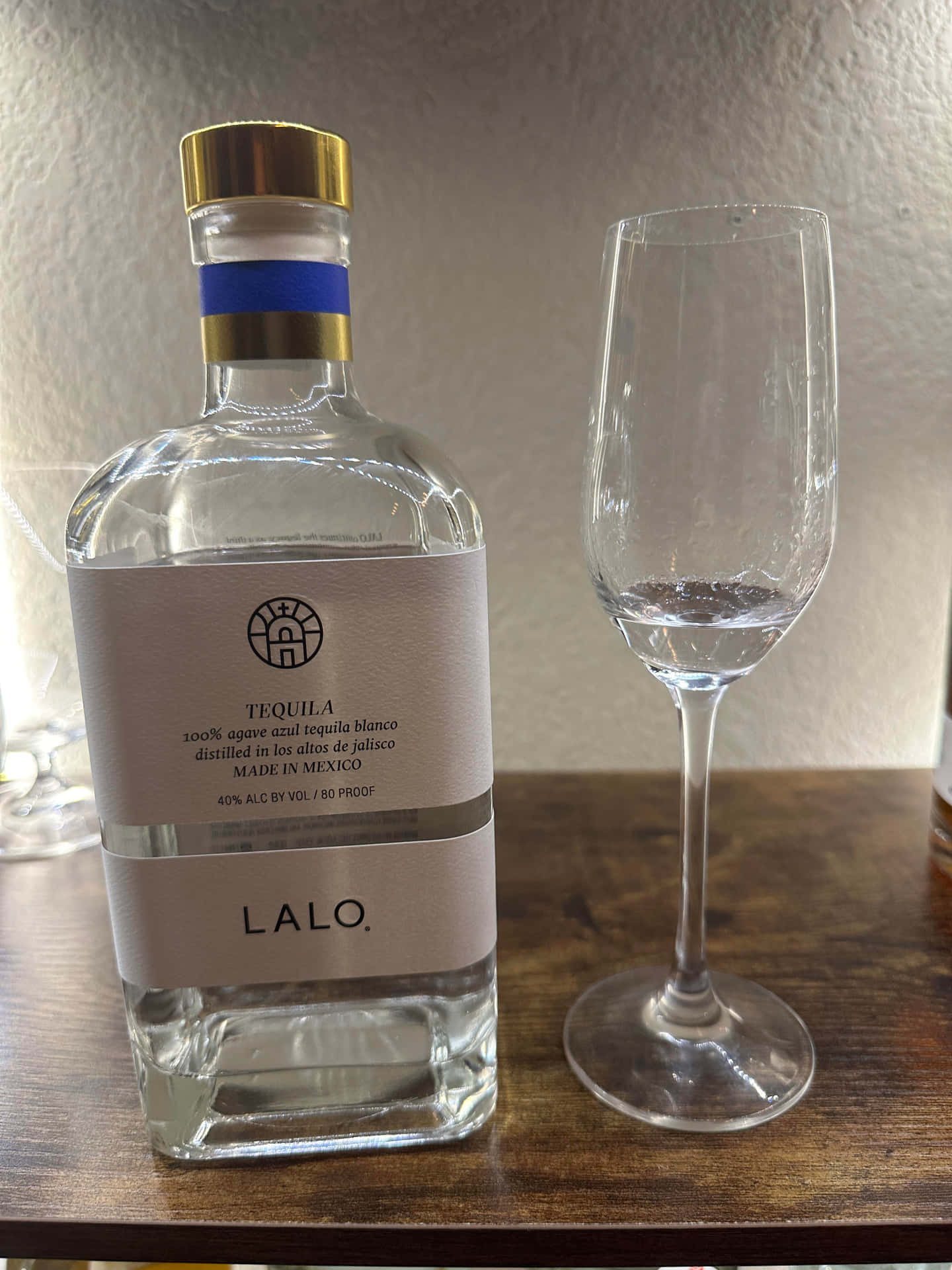  壁紙 — 一個透明的Lalo龍舌蘭酒瓶，金色瓶蓋，放在木桌上一個空的酒杯旁邊。