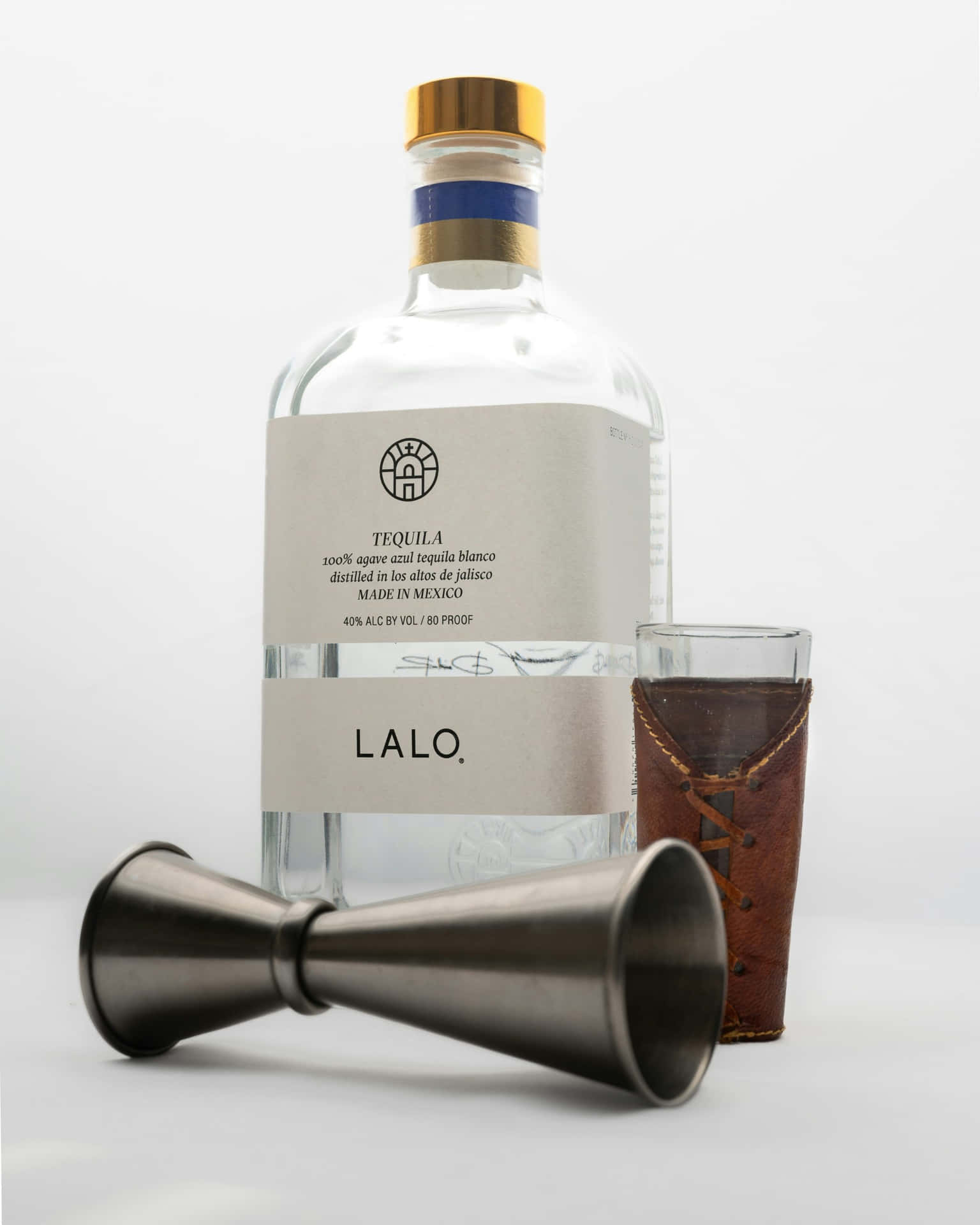 壁紙 — 一個透明的Lalo龍舌蘭酒瓶，金色瓶蓋和簡約標籤，旁邊放著一個皮革包裹的杯子和金屬量酒器在白色表面上。
