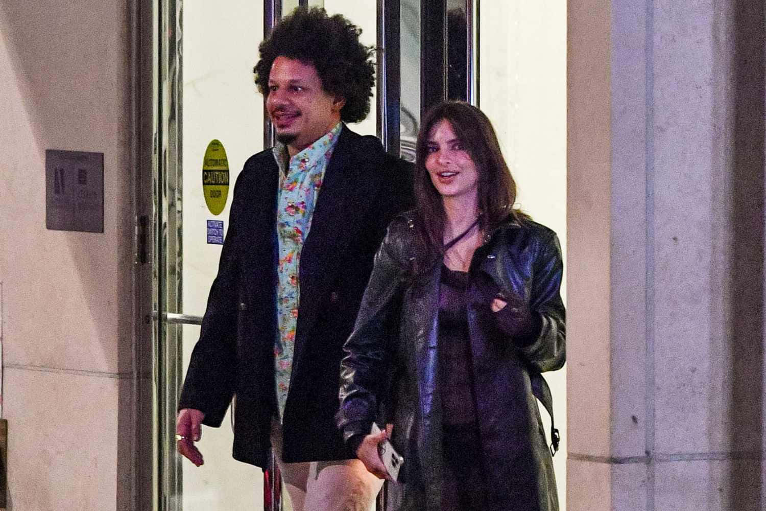  壁紙 — 一個微笑的捲髮男子和一個穿皮夾克的女子在夜晚的人造燈光下離開建築物。