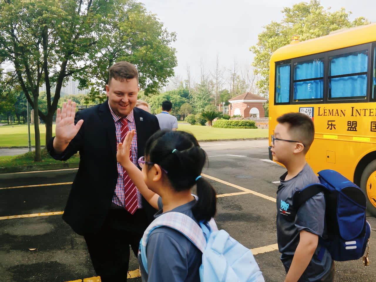  壁紙 — 一個微笑的西裝男子在綠樹成蔭的校園道路上，向一輛黃色校車附近的兩名學童揮手告別。