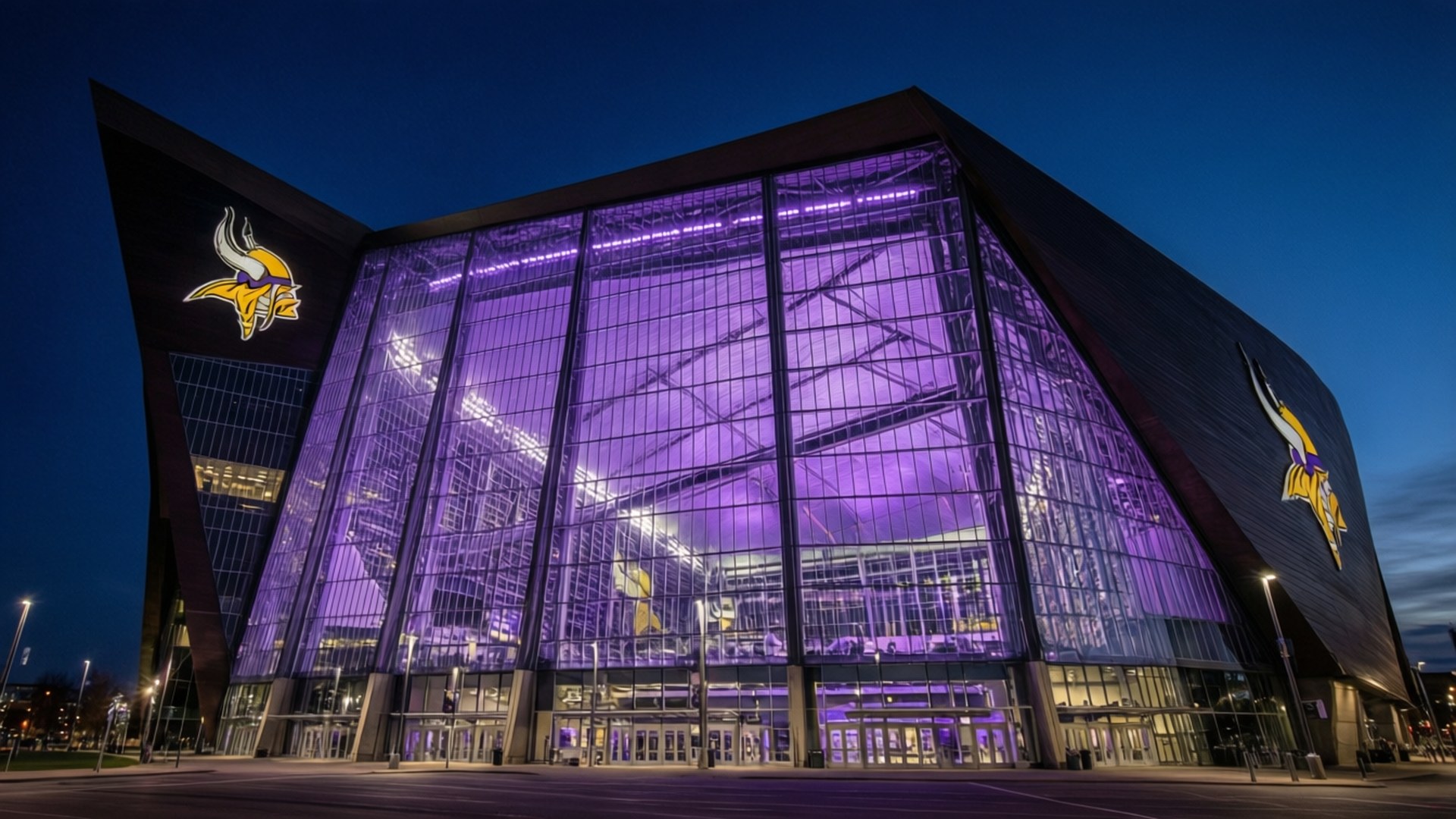 Vikings Stadium at Night 壁紙 — 一個現代玻璃體育場在深藍色的暮色天空中散發著充滿活力的紫色光芒。