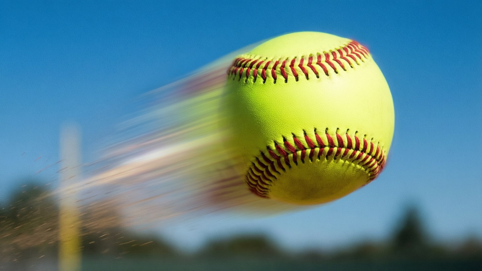 Soaring Softball 壁紙 — 一個鮮黃色的壘球帶著紅色縫線，在清晰的藍天背景下模糊地穿過空氣。