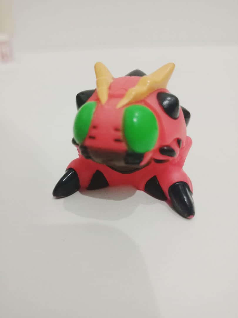 Digimon Tentomon 桌布 壁紙 — 一個小小的紅色塑膠玩具怪獸，有綠色眼睛、黑色爪子和黃色角，坐在一個純白色的表面上。