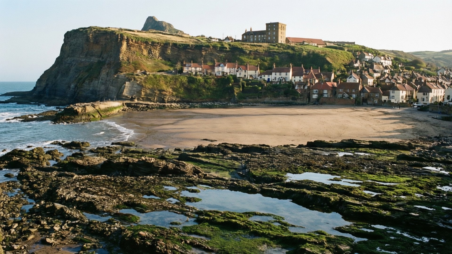 Robin Hood's Bay Tide 壁紙 — 一個沿海村莊依附在長滿草的懸崖上，俯瞰沙灘和有潮汐池的岩石海岸線。