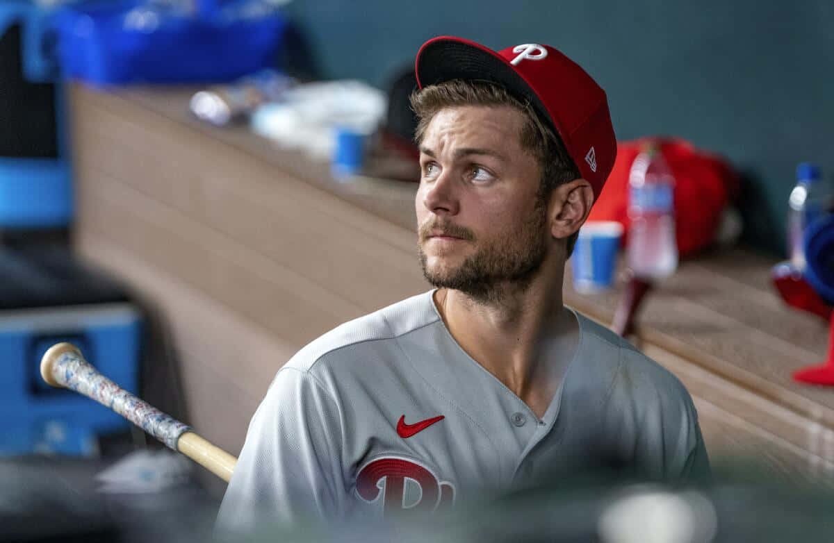 Trea Turner Phillies 桌布 壁紙 — 一個專注的棒球運動員，戴著紅色帽子，穿著灰色制服，坐在球員休息區，握著球棒，堅定地向上看。