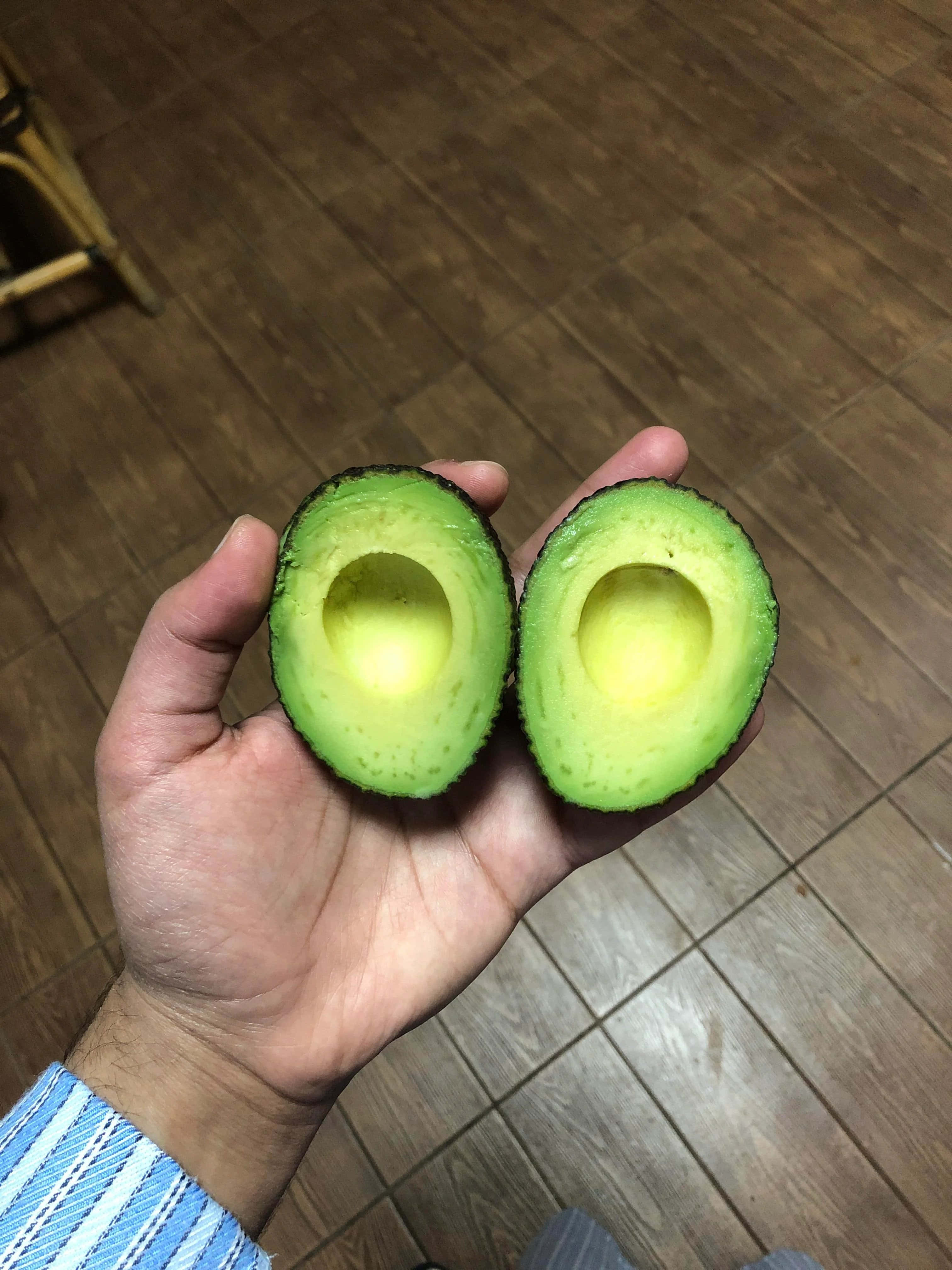 Avocado 桌布 壁紙 — 一隻手拿著兩個半邊成熟的酪梨，在木地板背景下露出奶油黃色的果肉。