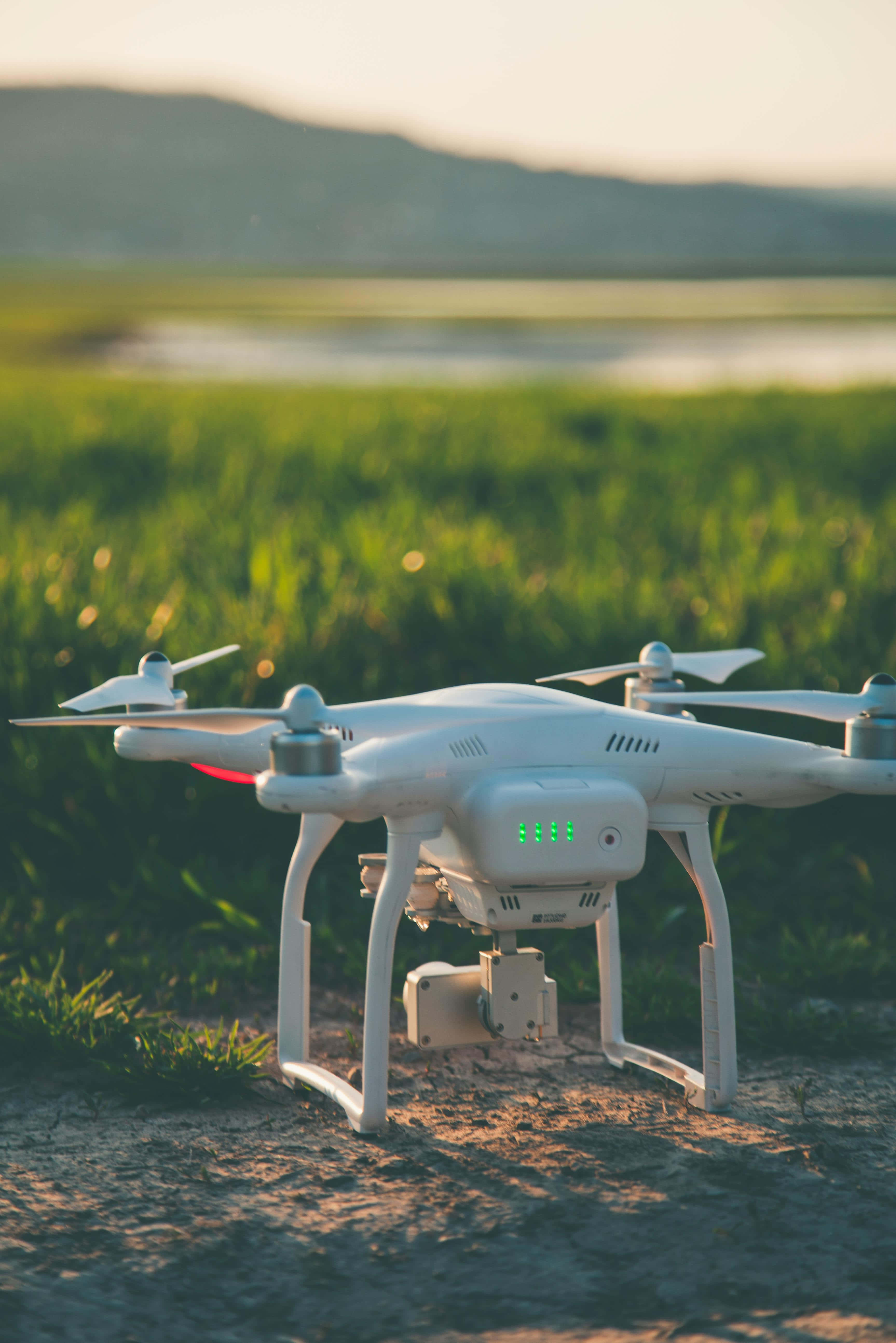 Drone Camera 桌布 壁紙 — 一架白色無人機帶有發光綠色燈光，在金色時刻停放在草地上，背景有山丘。