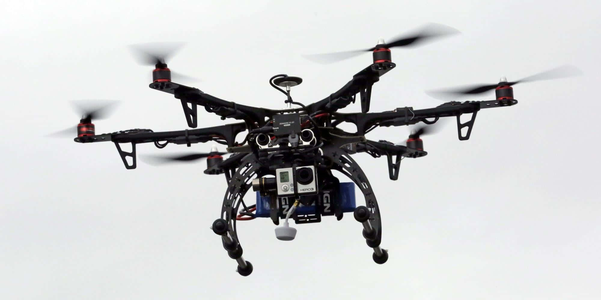 Drone Camera 桌布 壁紙 — 一架帶有紅色點綴的黑色六軸無人機，搭載攝像頭和傳感器，在蒼白的天空中盤旋。
