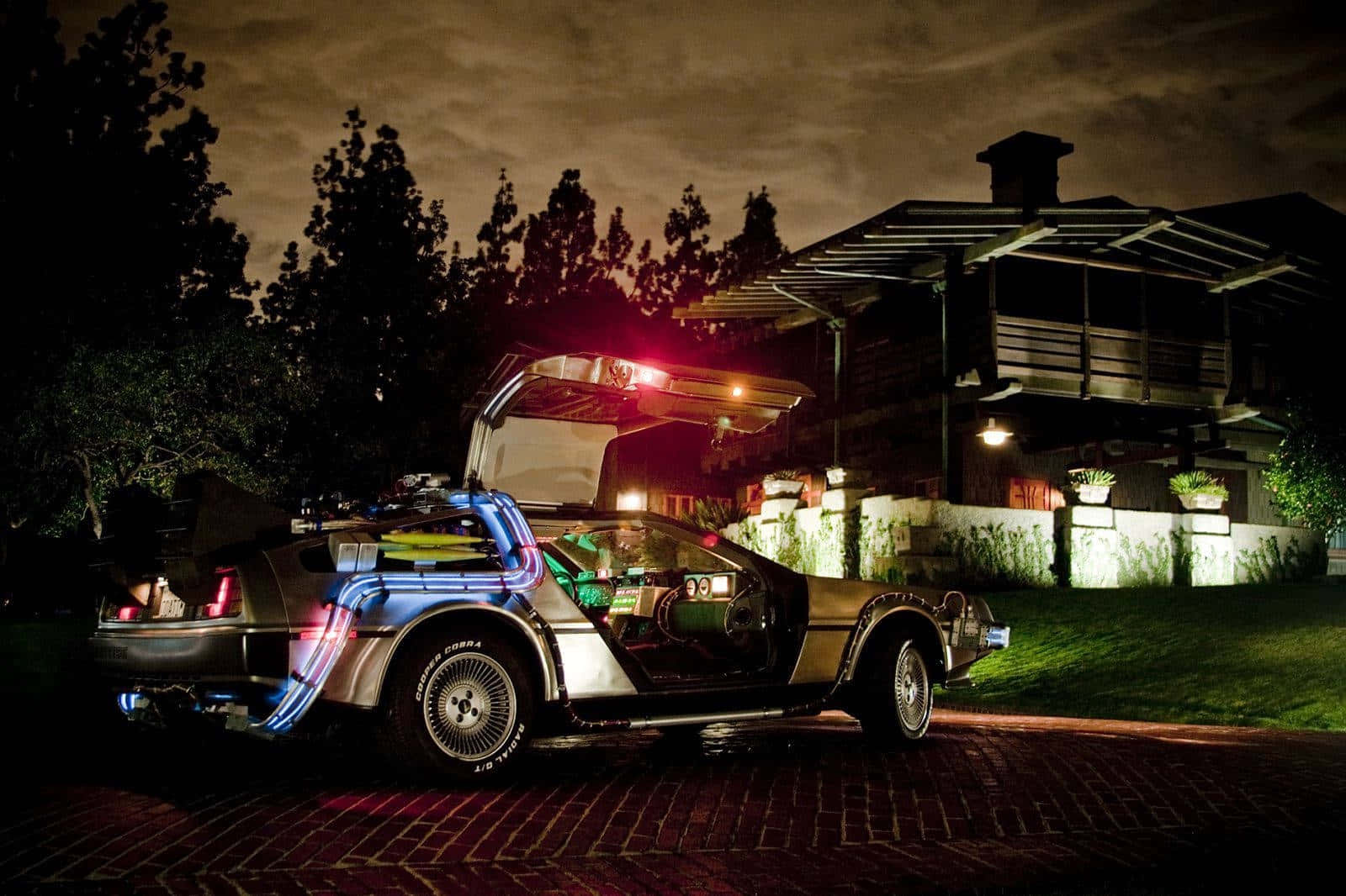  壁紙 — 一架發光的 DeLorean 時光機，打開鷗翼車門，夜晚停泊在磚砌車道上，天空多雲。