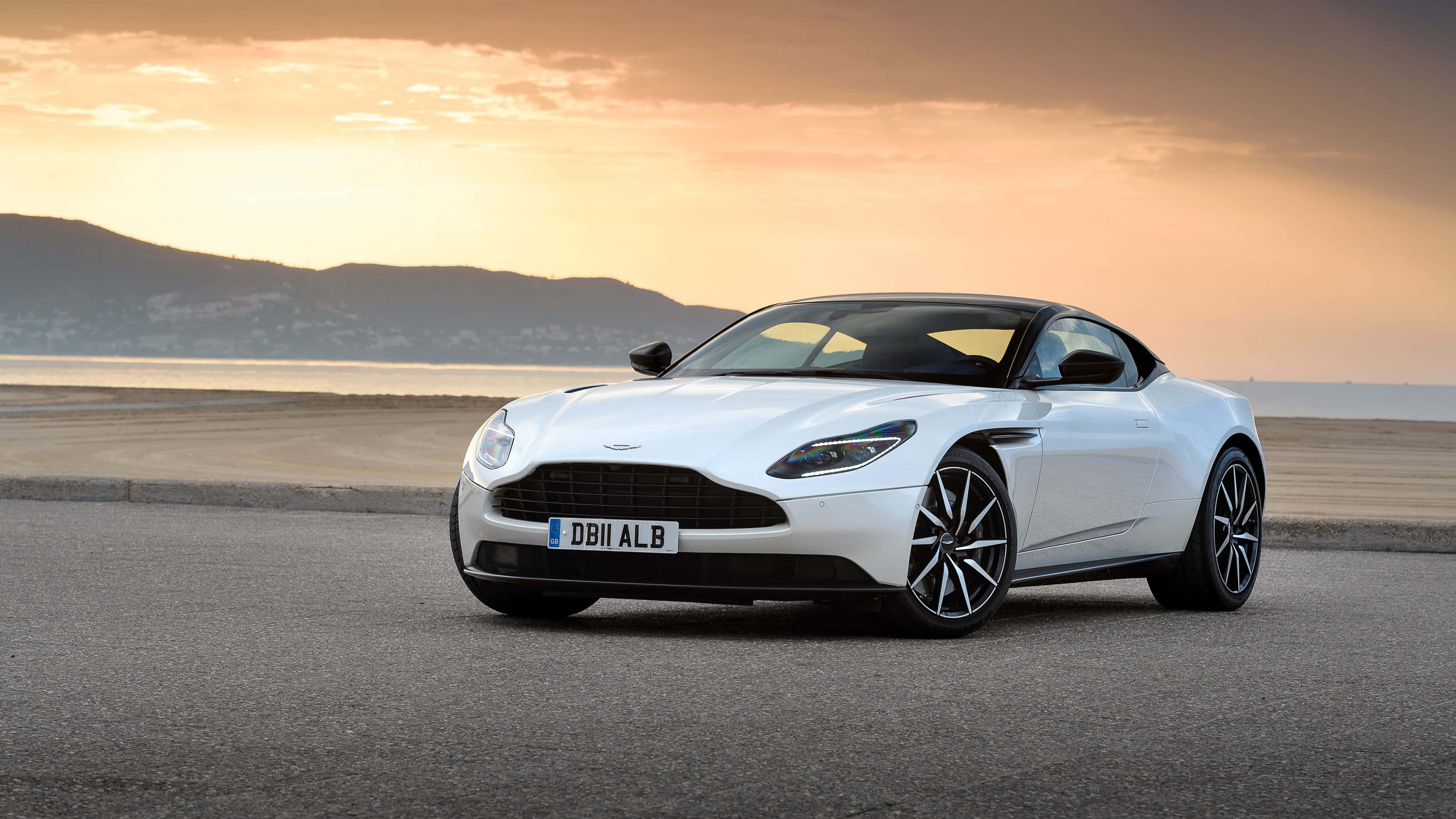 Aston Martin Db11 桌布 壁紙 — 一架流線型白色Aston Martin DB11停泊在日落時分的沿海公路上，背景有山脈和海洋。