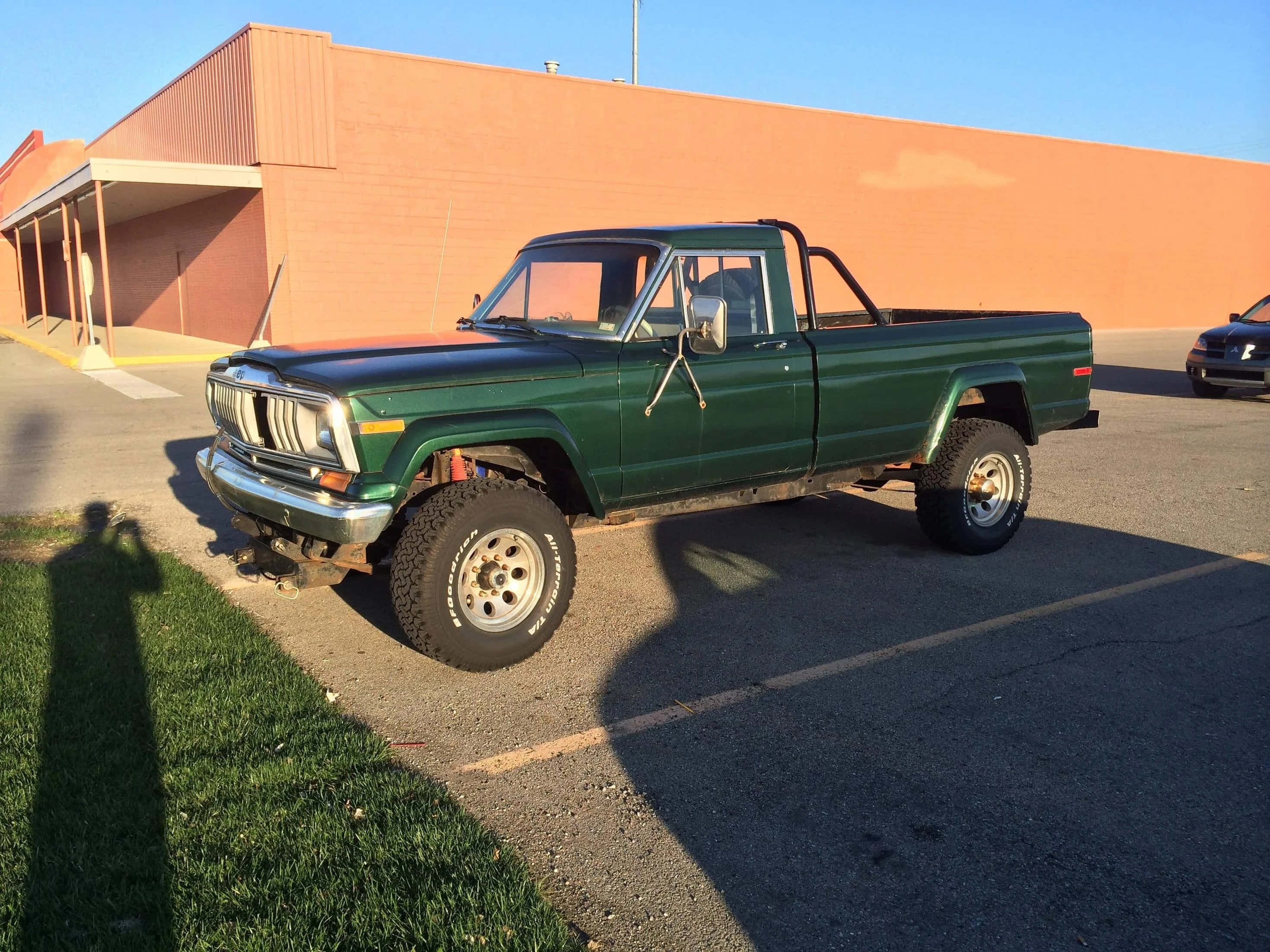 Jeep Comanche 桌布 壁紙 — 一架升高了的綠色經典皮卡車停放在一個停車場，旁邊是一棟米色建築，天空晴朗蔚藍。