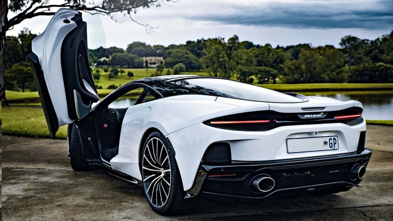 Mclaren Gt 桌布 壁紙 — 一架時尚的白色McLaren跑車，蝴蝶門打開，停泊在池塘旁邊的車道上，天空多雲。