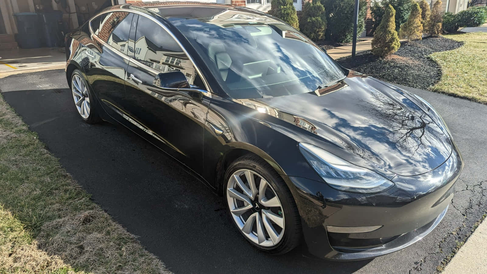 Tesla Model 3 桌布 壁紙 — 一架時尚的黑色Tesla Model 3停放在住宅車道上，反射出晴朗的天空和光禿的樹木。