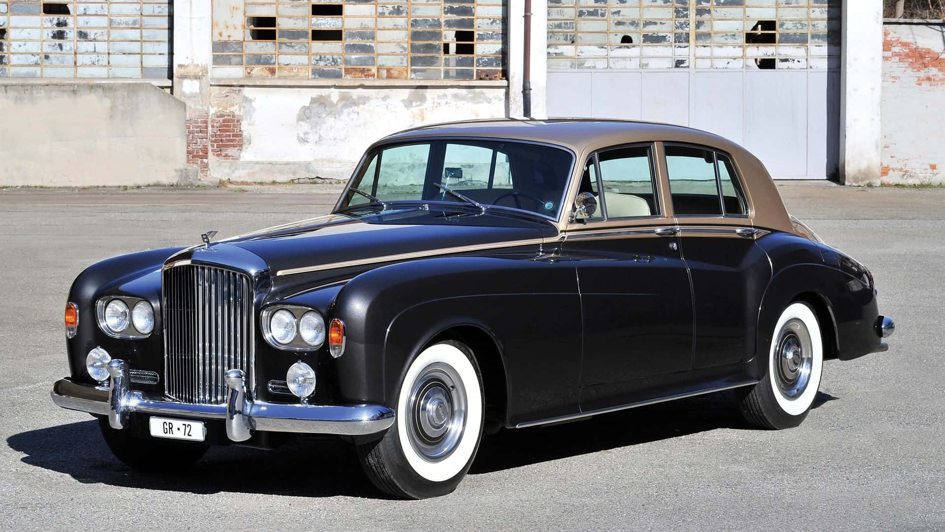 Bentley S3 桌布 壁紙 — 一架時尚的黑色 vintage Bentley 配備白色牆壁輪胎，停泊在風化的工業建築前。