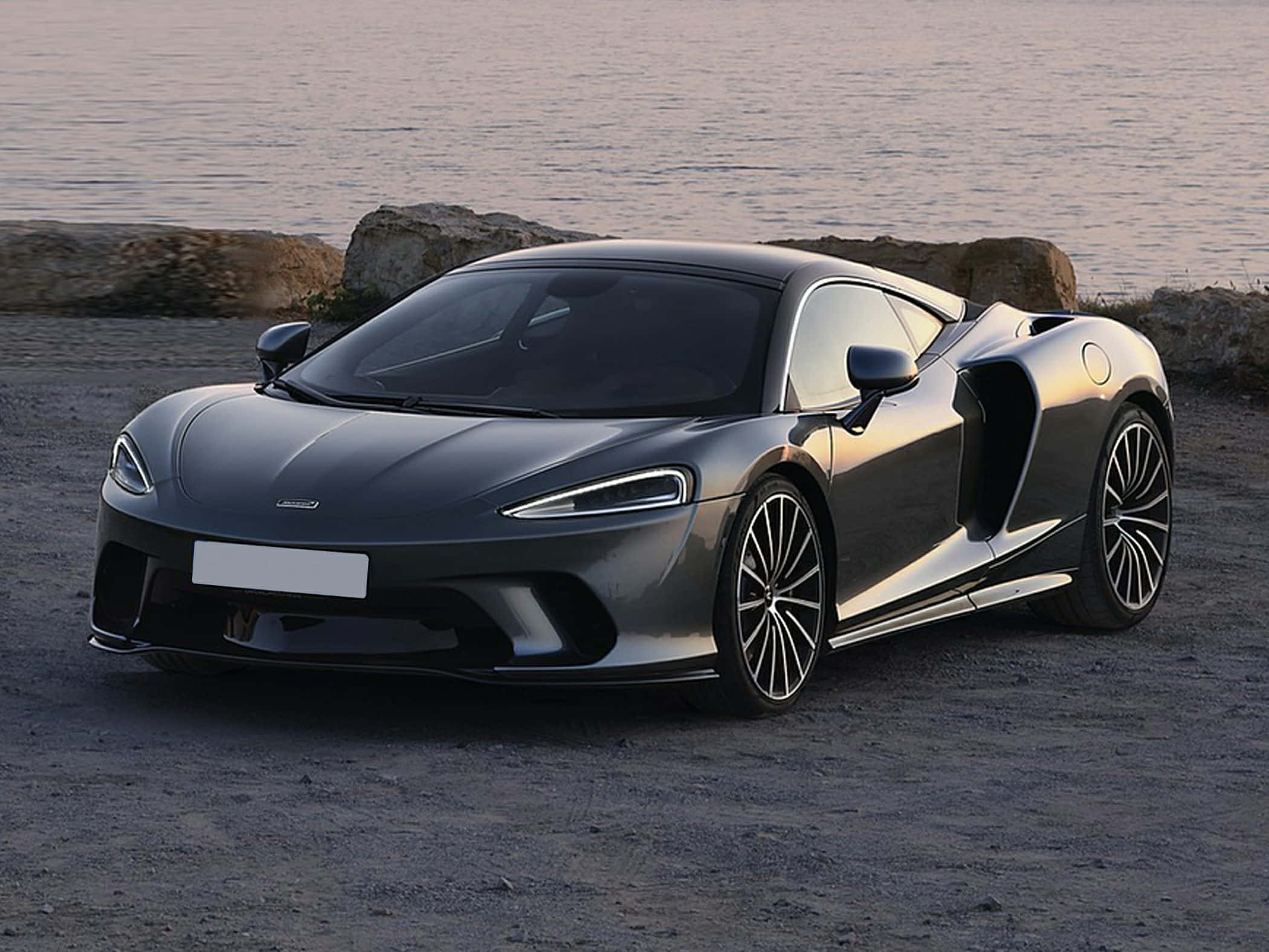 Mclaren Gt 桌布 壁紙 — 一架時尚的灰色McLaren跑車停泊在日落時分的岩石海岸上，背後是平靜的水面。
