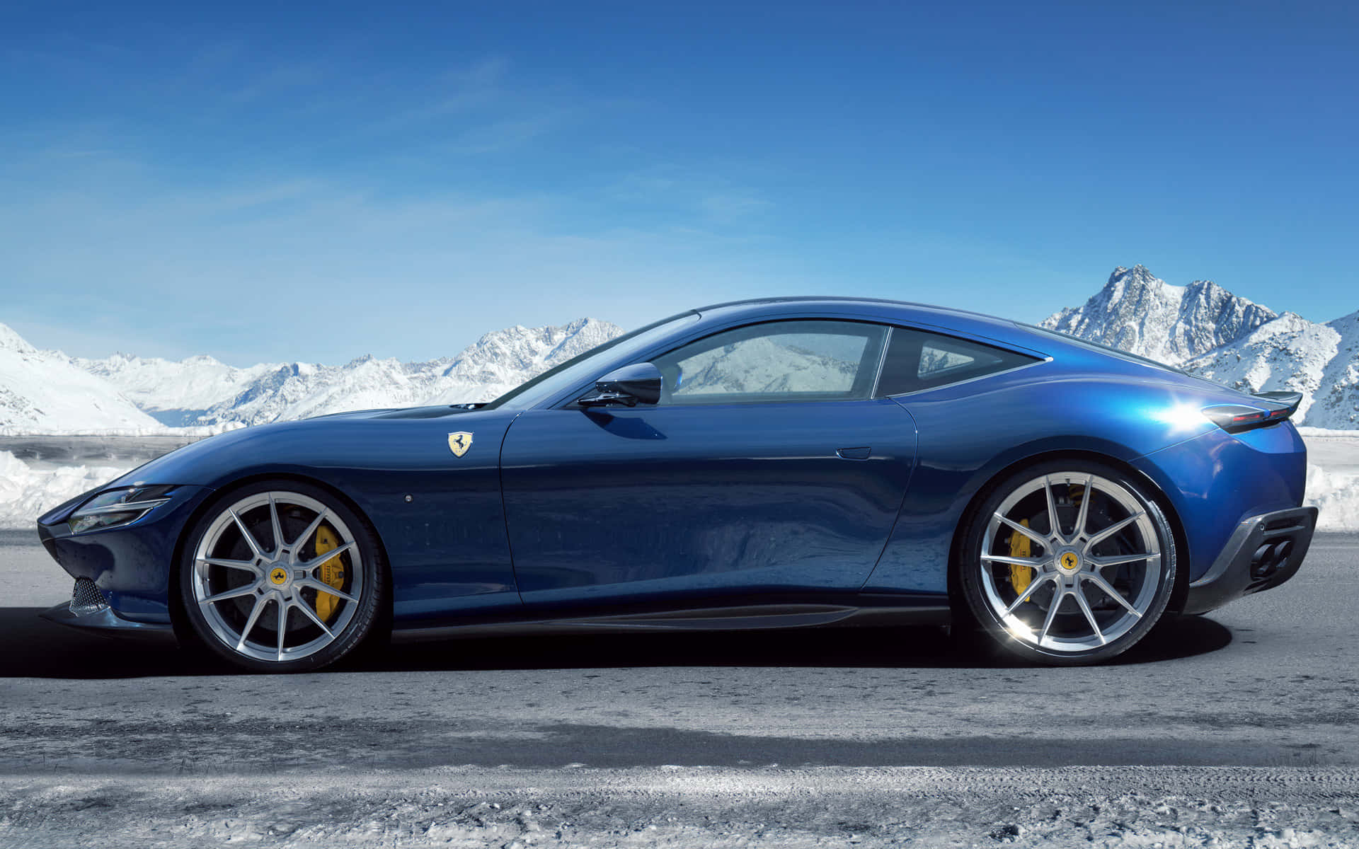 Ferrari Roma 桌布 壁紙 — 一架時尚的藍色法拉利跑車在明亮的陽光下閃閃發光，背景是白雪覆蓋的山脈。