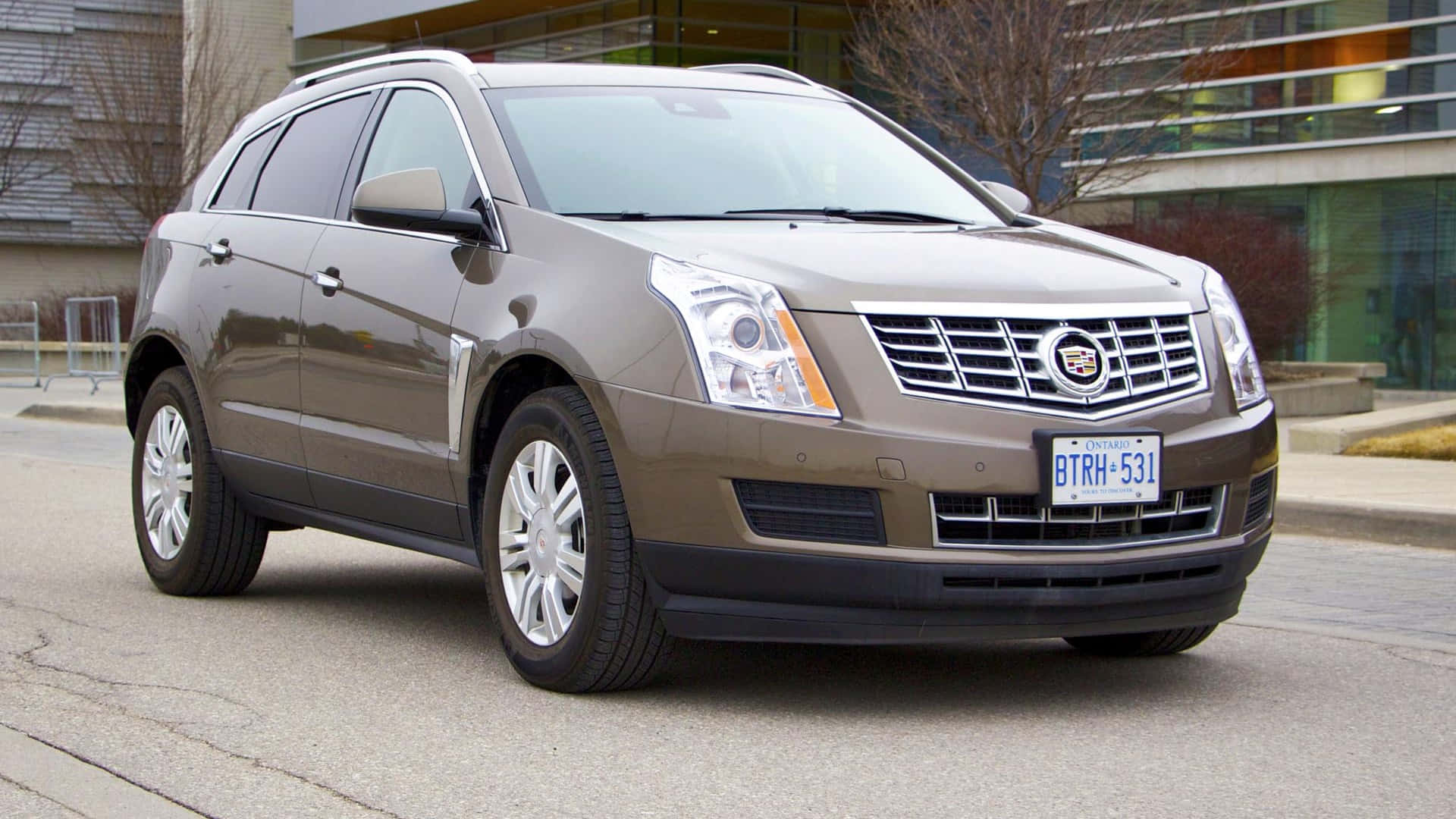 Cadillac Srx 桌布 壁紙 — 一架時尚的青銅色Cadillac SRX停放在城市人行道上，背景是現代建築和光禿禿的樹木。
