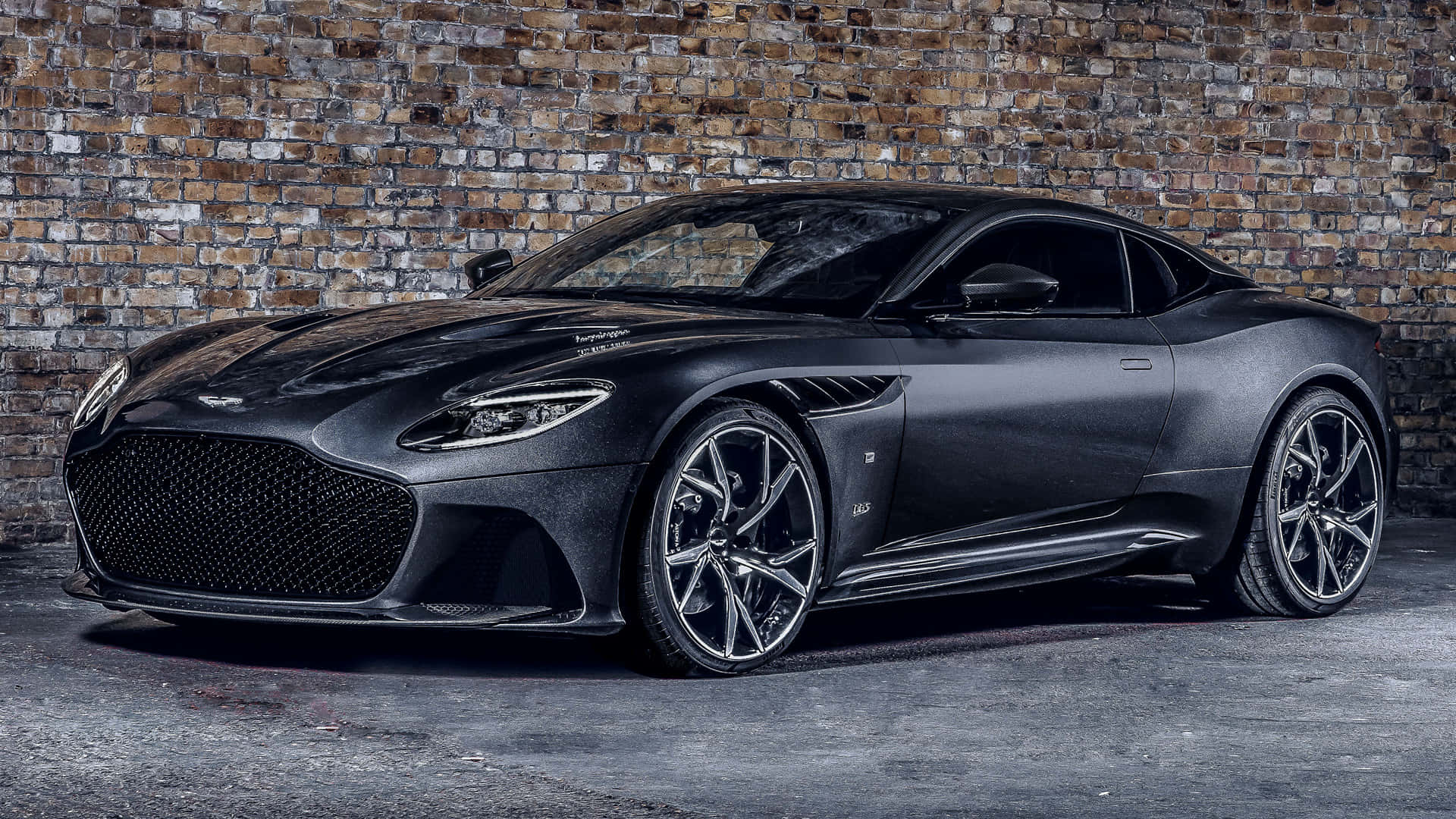 Aston Martin Dbs Superleggera 桌布 壁紙 — 一架時尚的啞光黑色 Aston Martin DBS Superleggera 停靠在昏暗車庫中風化的磚牆旁。