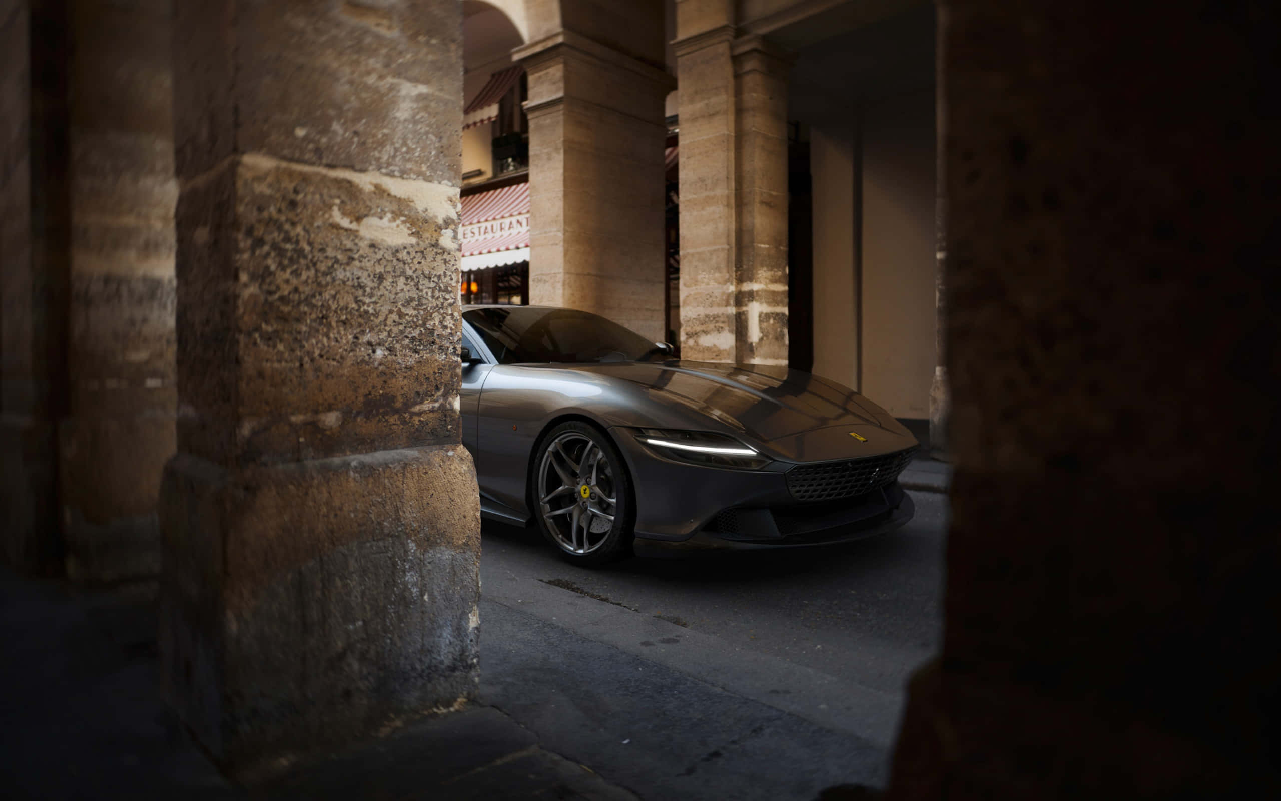 Ferrari Roma 桌布 壁紙 — 一架時尚的銀色法拉利停泊在昏暗城市庭院中的古老石拱門下。