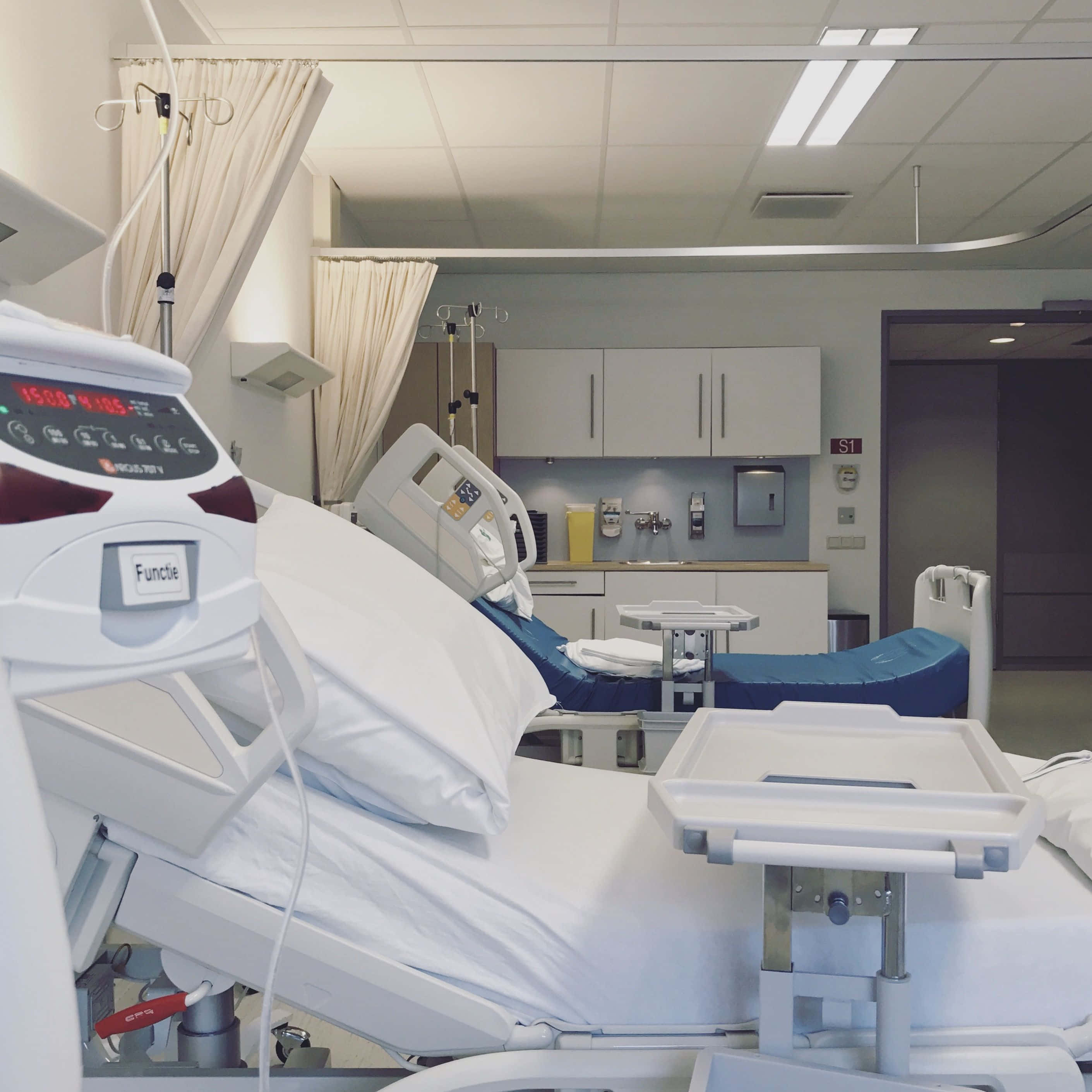 Hospital Bed 桌布 壁紙 — 一間乾淨現代化的醫院病房，設有兩張可調節病床、醫療設備和白色窗簾，在明亮的螢光燈下。