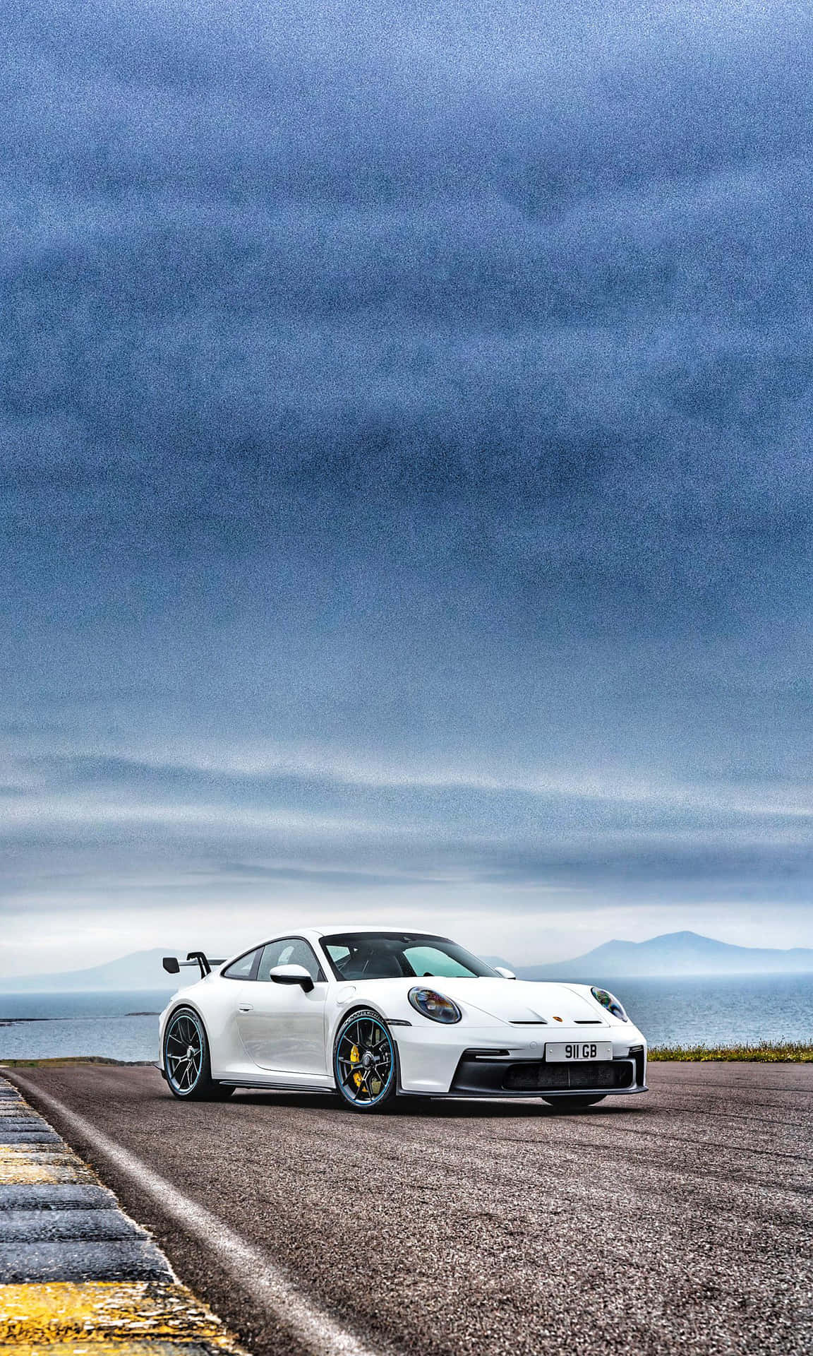  壁紙 — 一輛白色保時捷911 GT3 RS停泊在沿海公路上，天空陰沉多變，遠方有山脈和海洋。