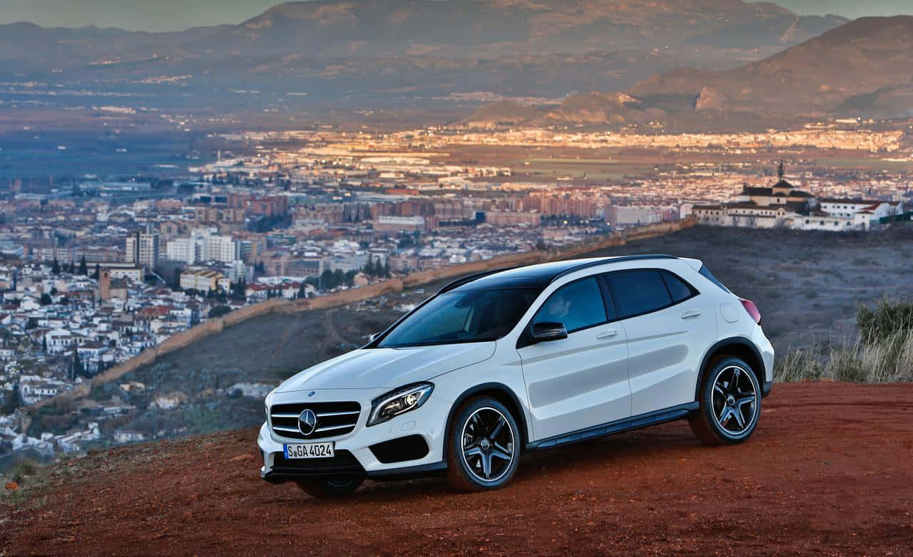 Mercedes Benz Gla-class 桌布 壁紙 — 一輛白色賓士GLA-Class SUV停放在紅色泥土觀景點上，俯瞰著沐浴在金色夕陽下的廣闊城市和山脈。