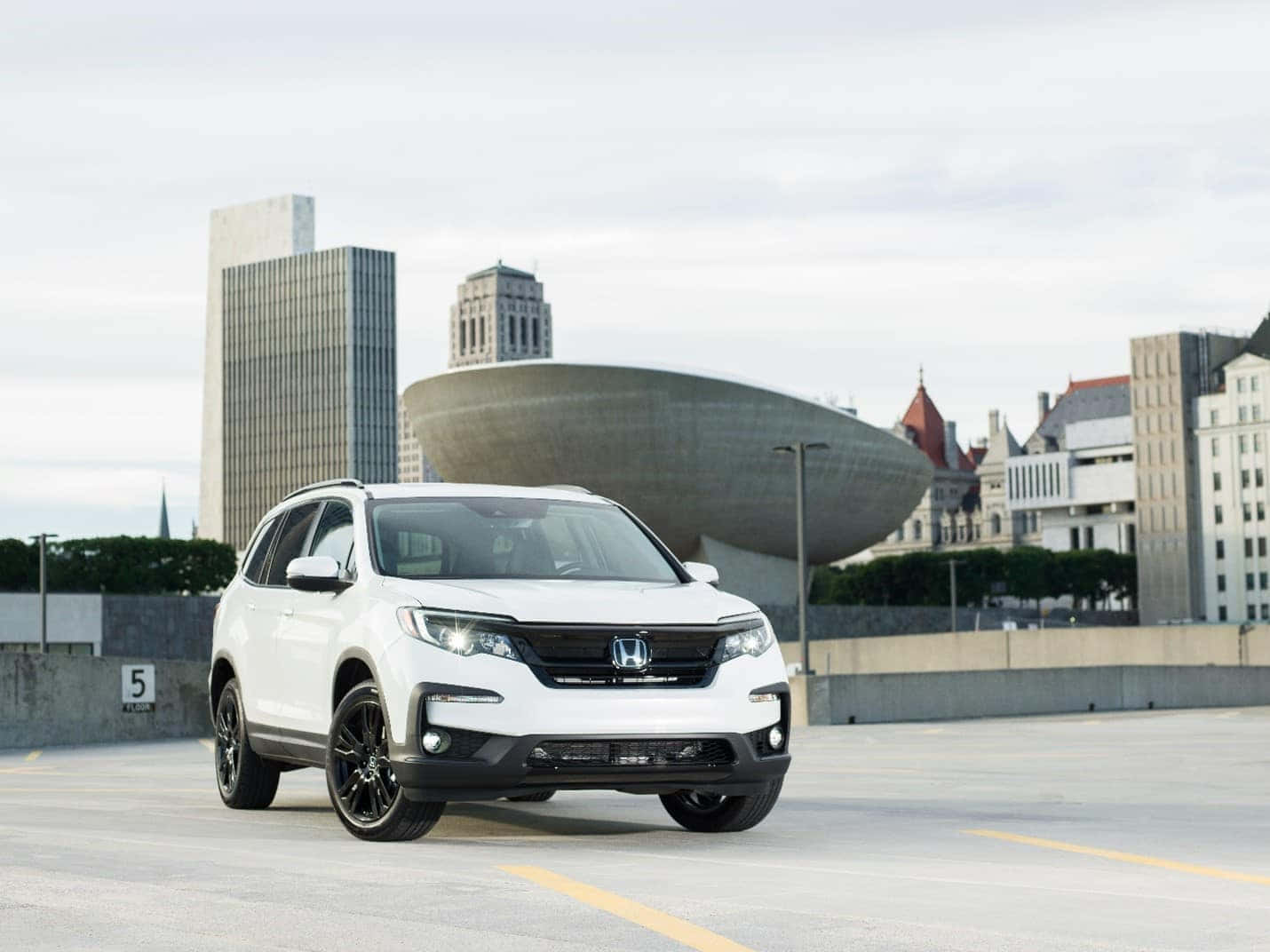 Honda Pilot 桌布 壁紙 — 一輛白色本田SUV配黑色輪圈，停放在現代建築背景的城市停車場。