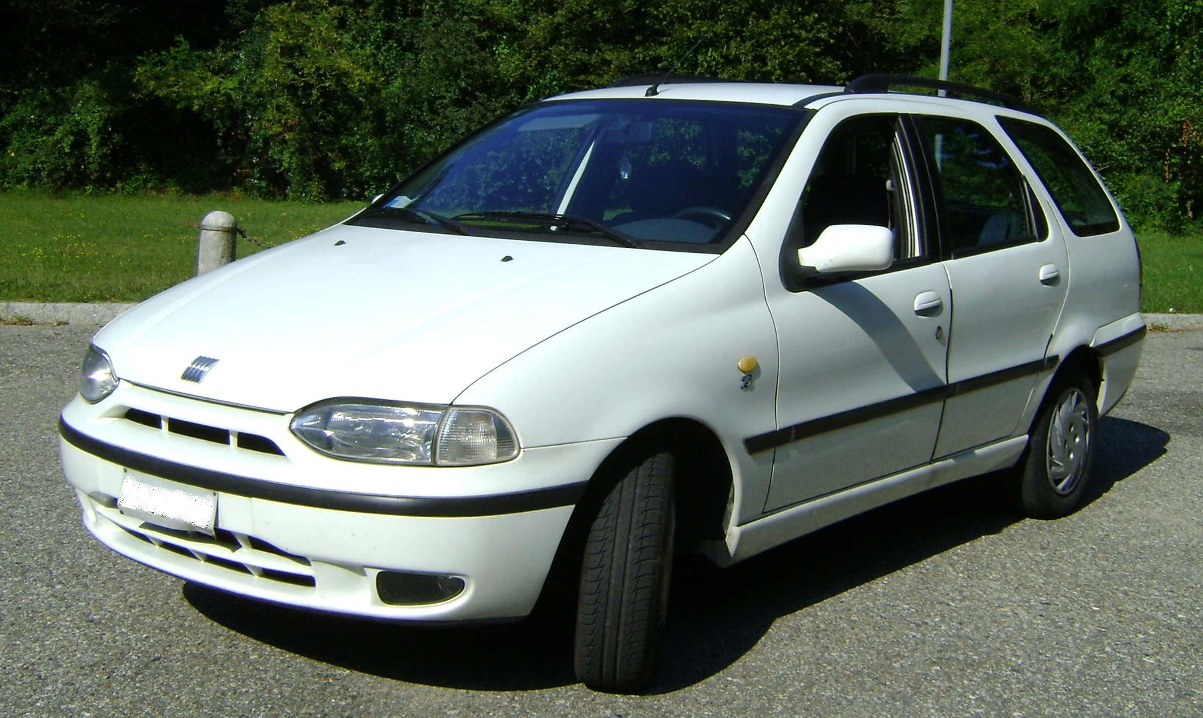 Fiat Palio 桌布 壁紙 — 一輛白色Fiat Stilo旅行車停在柏油路上，背景有綠樹和草地，在明亮的日光下。