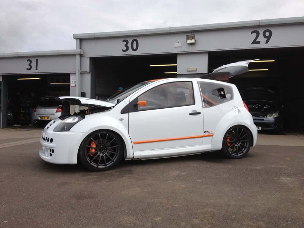 Citroen C2 Super 1600 桌布 壁紙 — 一輛白色改裝的雷諾Clio，帶有橙色點綴和黑色車輪，停泊在帶有編號的車庫位外，天空多雲。