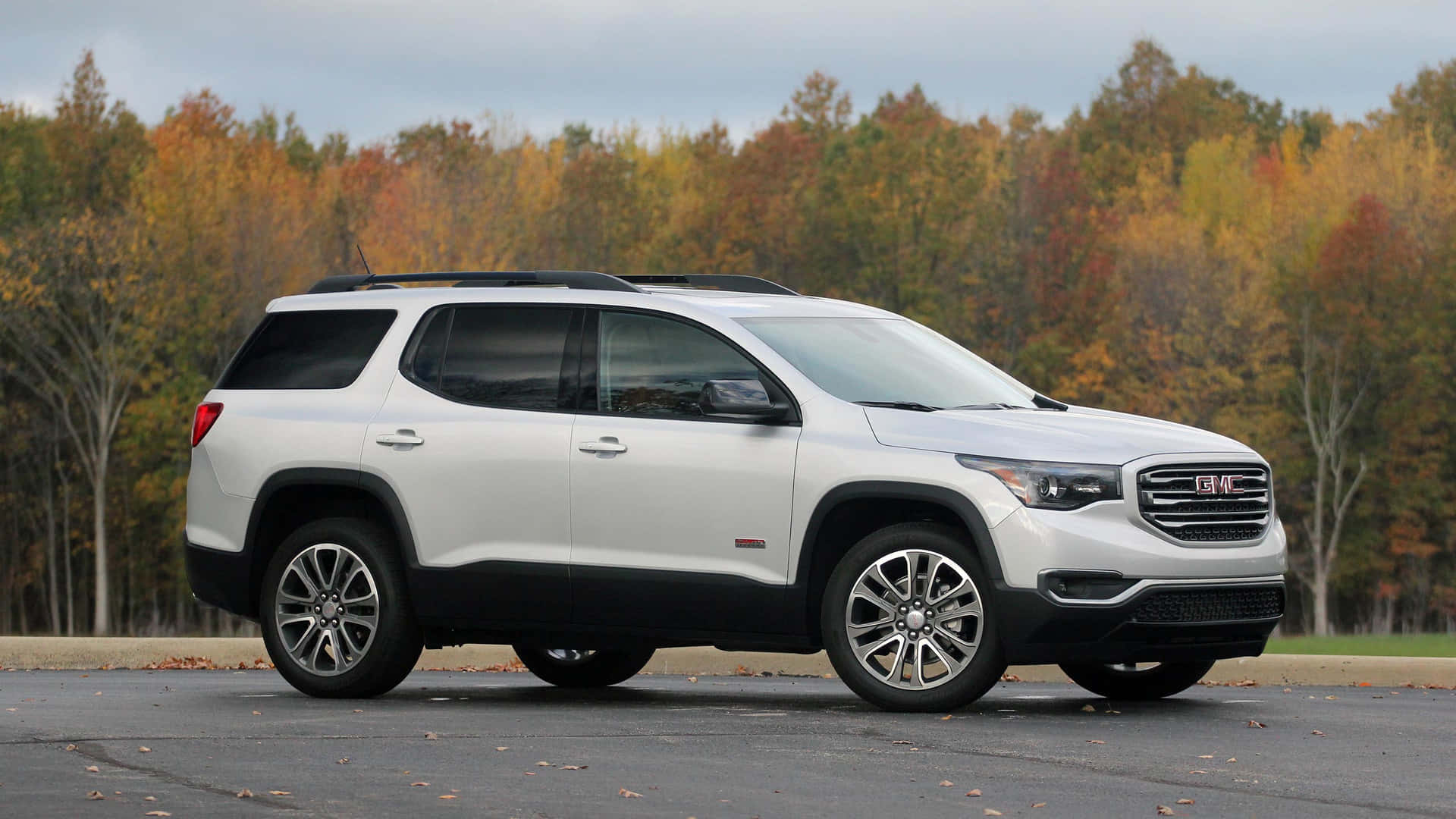Gmc Acadia 桌布 壁紙 — 一輛白色GMC Acadia SUV停在瀝青路上，背景有秋天的樹木，展示橙色、黃色和紅色的樹葉，天空多雲。