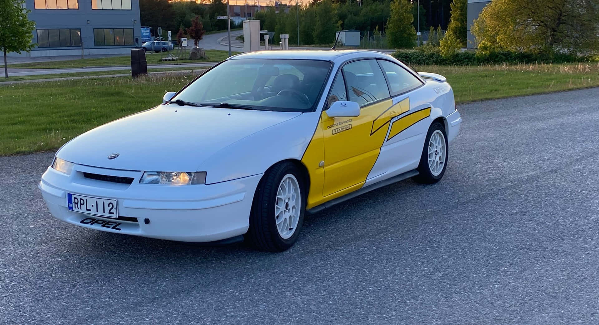 Opel Calibra 桌布 壁紙 — 一輛白色和黃色的歐寶運動型轎跑車停放在日落時分的碎石路上，背景有建築物和樹木。