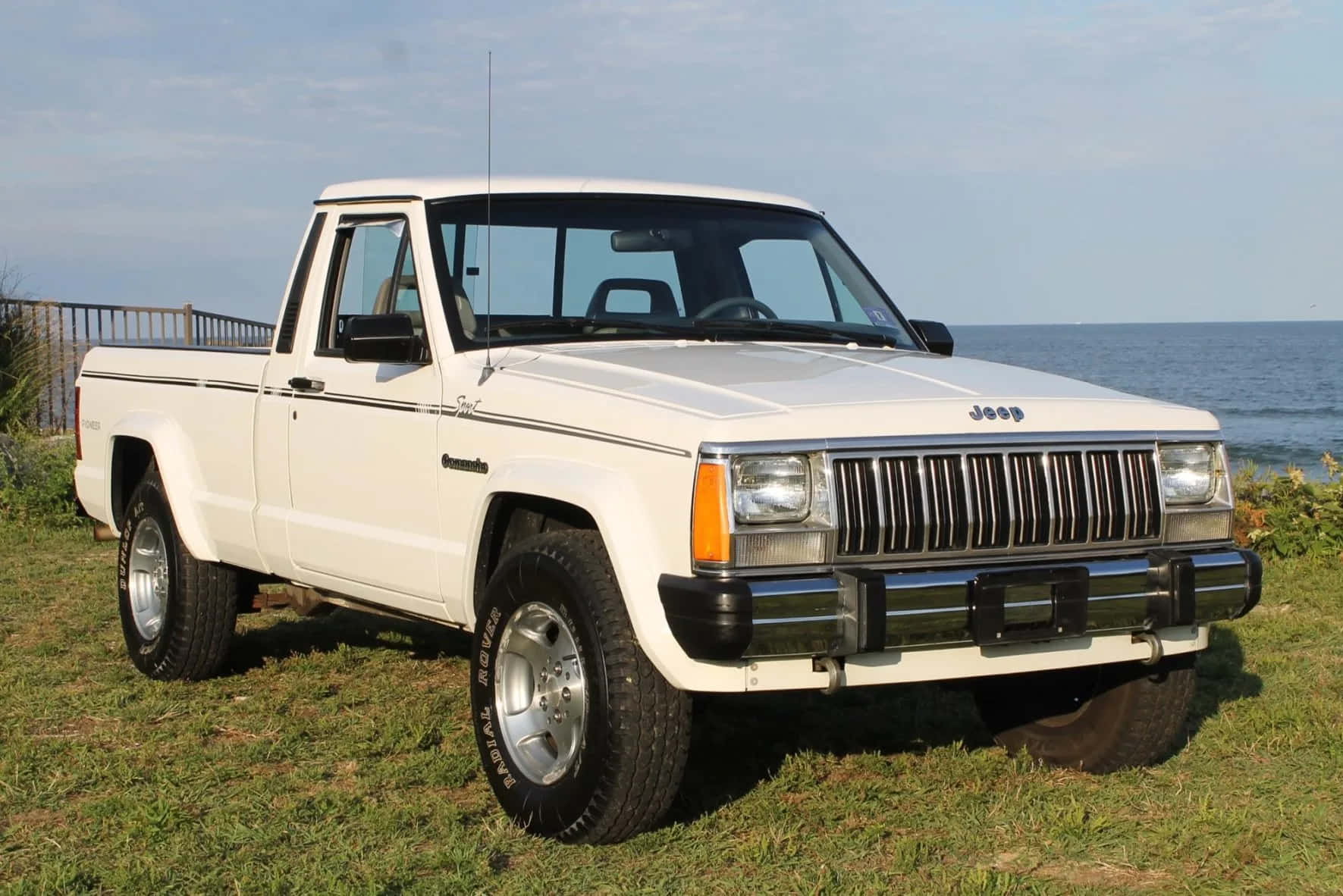 Jeep Comanche 桌布 壁紙 — 一輛白色Jeep Comanche皮卡車停在靠近海洋的草地上，天空晴朗。