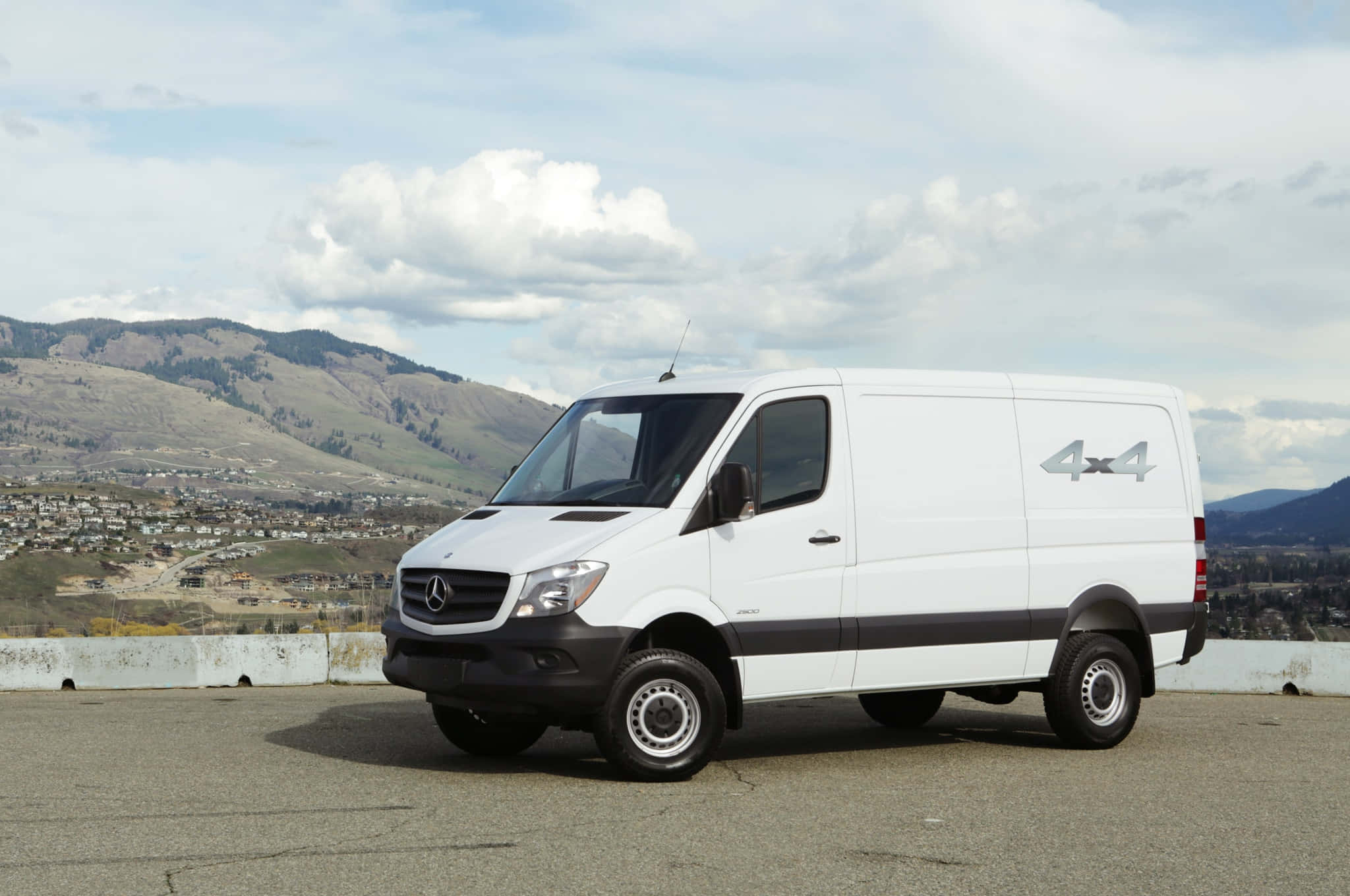 Mercedes Benz Sprinter 桌布 壁紙 — 一輛白色 Mercedes-Benz Sprinter 4x4 廂型車停放在鋪砌的觀景點，下方是起伏的丘陵和城鎮，天空部分多雲。