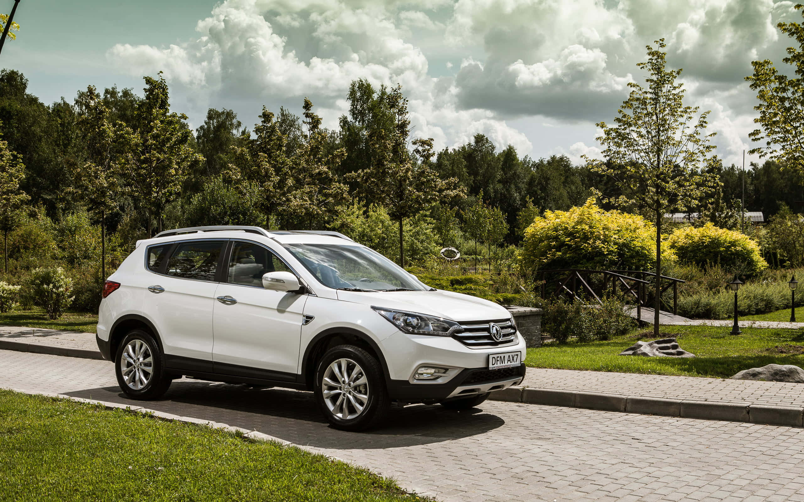 Dongfeng 桌布 壁紙 — 一輛白色SUV停放在鋪砌小徑上，旁邊有綠草和樹木，天空多雲。