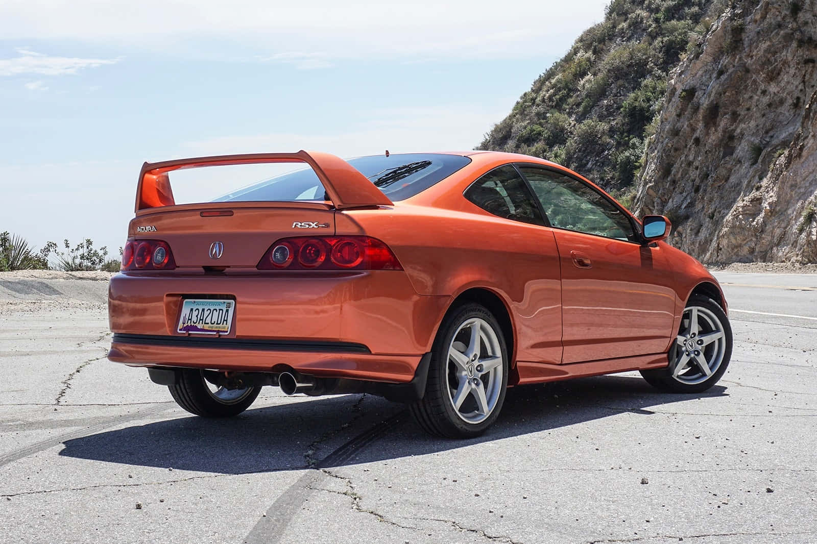 Acura Rsx 桌布 壁紙 — 一輛充滿活力的橙色Acura RSX Type-S雙門跑車停放在裂開的柏油路上，旁邊是岩石山丘，在清澈的藍天下。