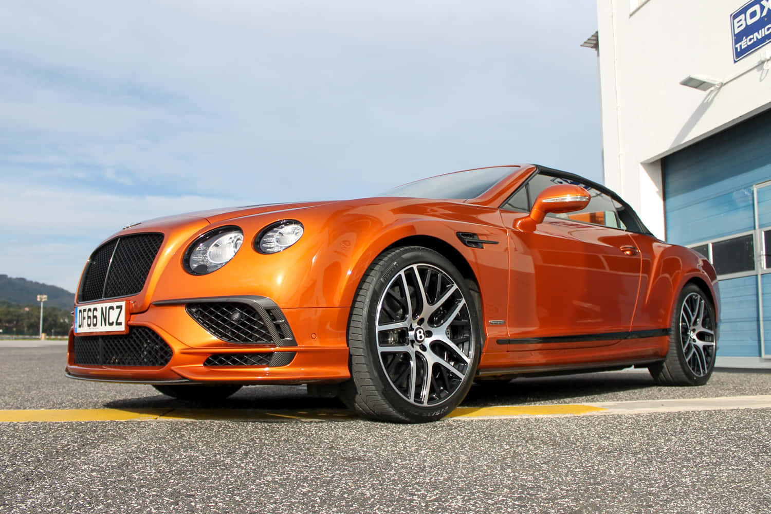 Bentley Continental Supersports 桌布 壁紙 — 一輛充滿活力的橙色賓利敞篷車停放在白色建築物旁邊的瀝青路面上，天空清澈湛藍。