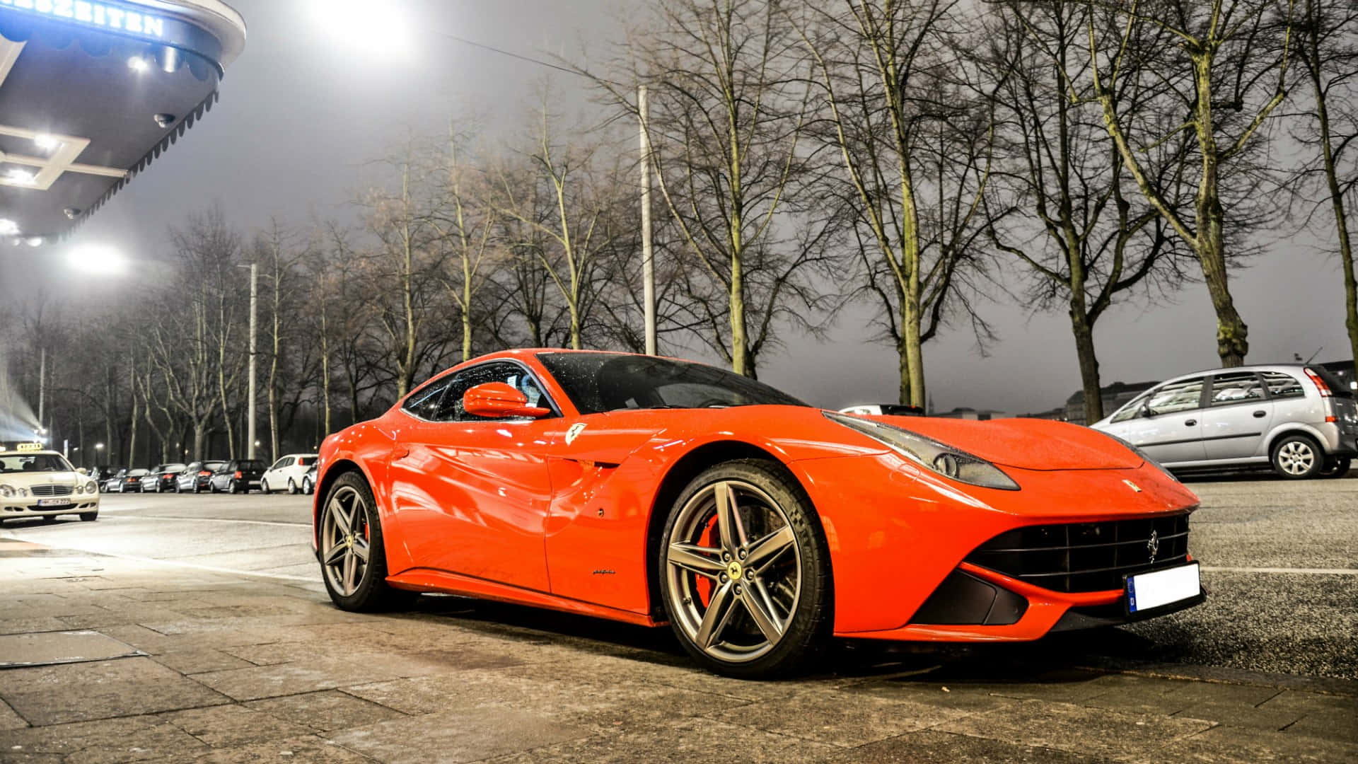 Ferrari F12 Berlinetta 桌布 壁紙 — 一輛充滿活力的橙色法拉利跑車在明亮的燈光下閃閃發光，停放在夜間城市停車場，周圍是光禿禿的樹木。