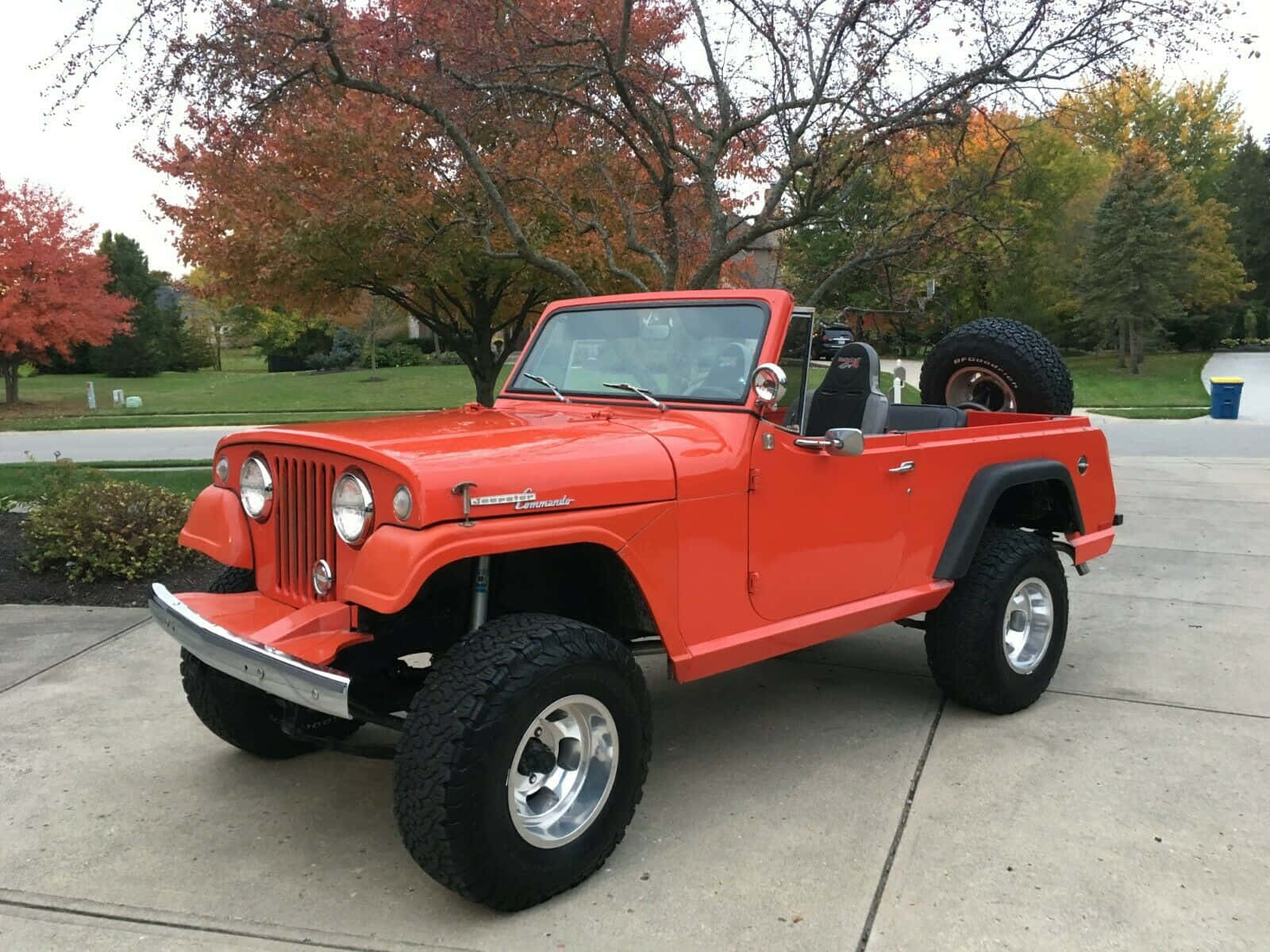 Jeepster 桌布 壁紙 — 一輛充滿活力的橙色經典Jeep Wrangler，配備大型輪胎，停在混凝土車道上，周圍是紅葉和橙葉的秋樹。