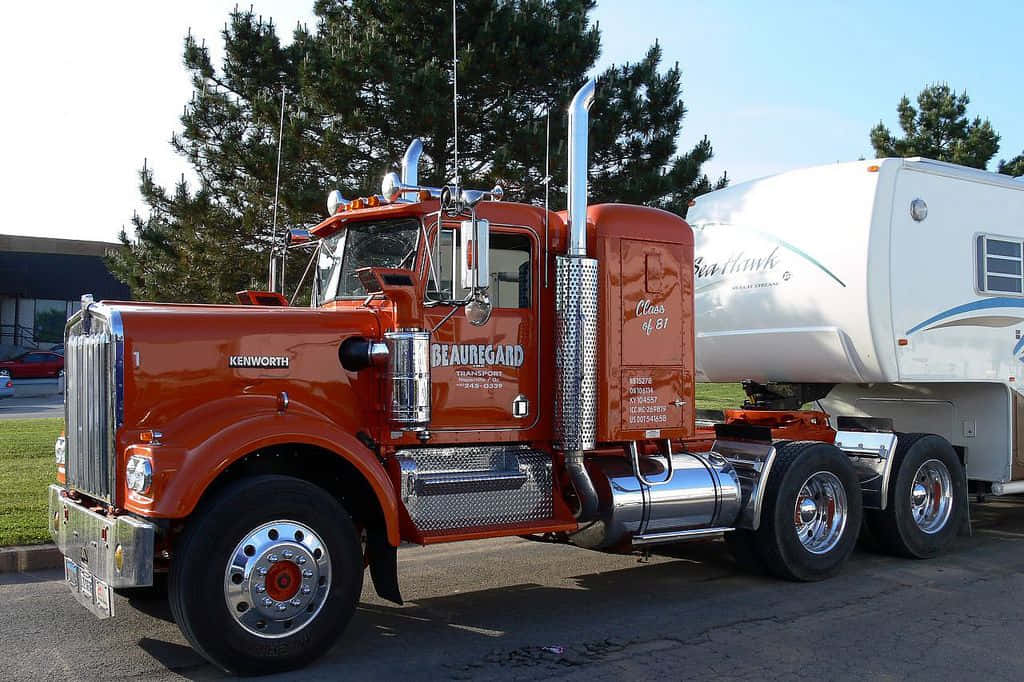 Kenworth C500 桌布 壁紙 — 一輛充滿活力的橙色Kenworth半掛卡車帶有鍍鉻裝飾，拖著一輛停在瀝青路面上的白色第五輪露營車，背景是晴朗的藍天。