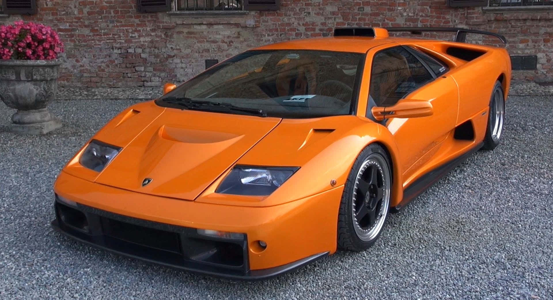 Lamborghini Diablo 桌布 壁紙 — 一輛充滿活力的橙色藍寶堅尼 Diablo SV 在日光下閃閃發光，停泊在磚牆和花盆旁邊的碎石上。