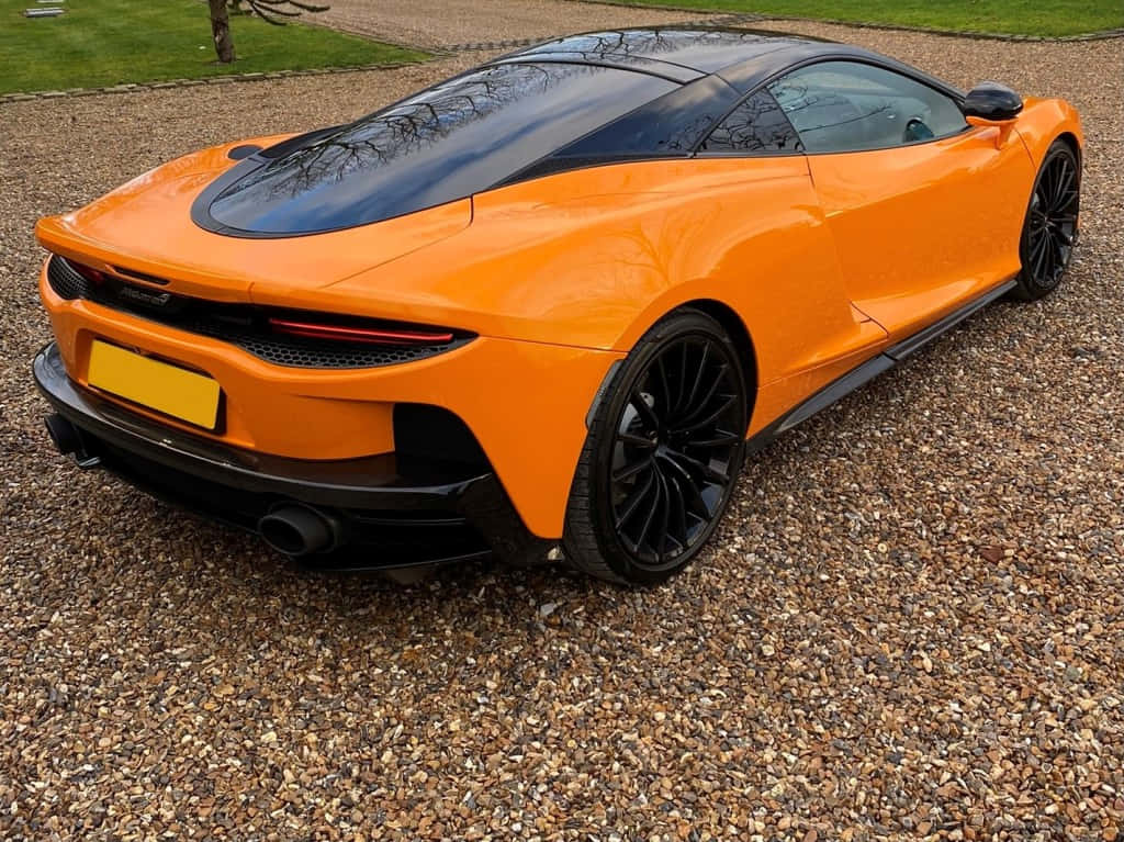 Mclaren Gt 桌布 壁紙 — 一輛充滿活力的橙色McLaren跑車，帶有黑色點綴，停在日光下的碎石車道上。