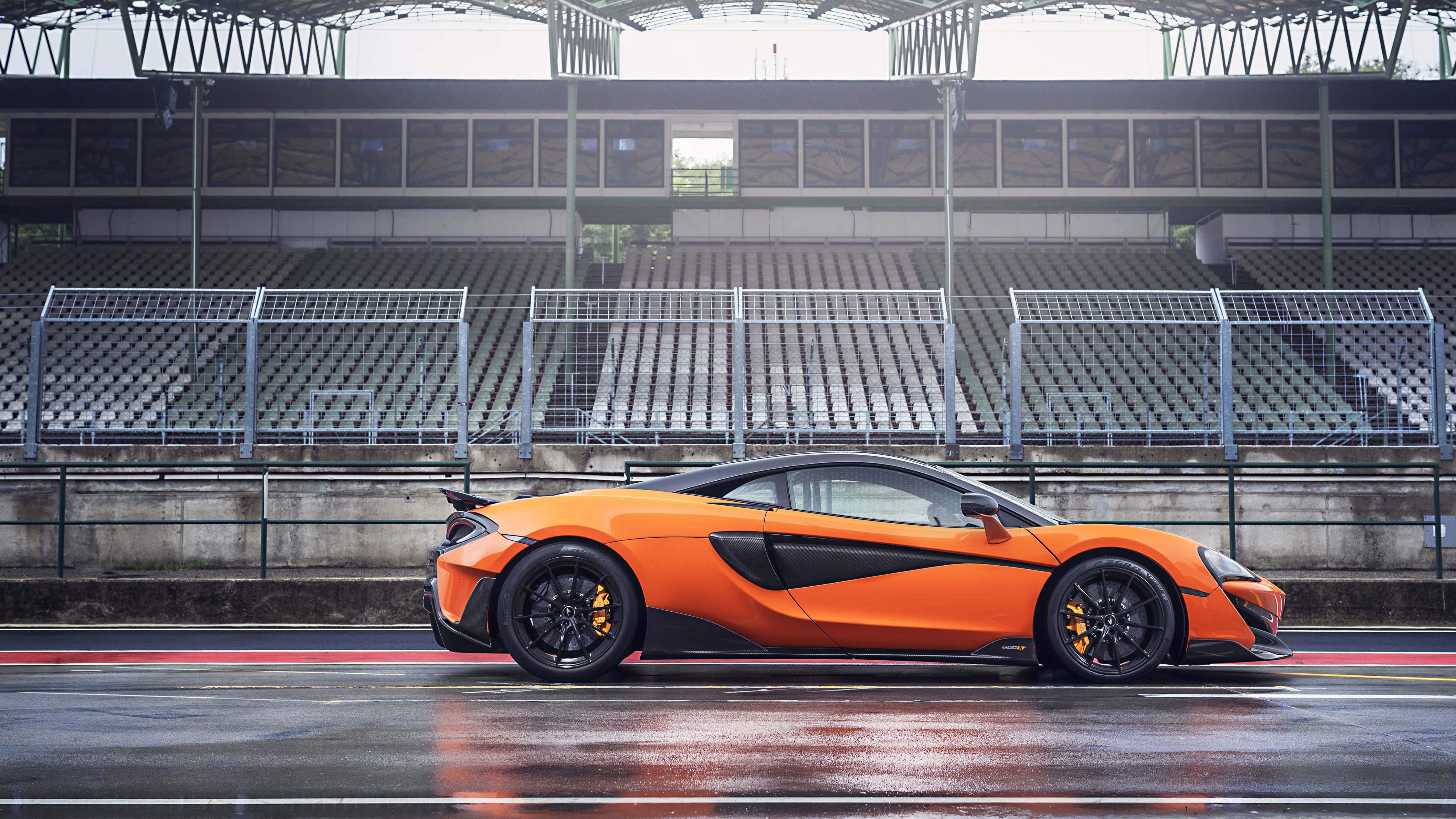 Mclaren 600lt 桌布 壁紙 — 一輛充滿活力的橙色McLaren跑車在潮濕的賽道上閃閃發光，天空多雲，後面是空無一人的看台。
