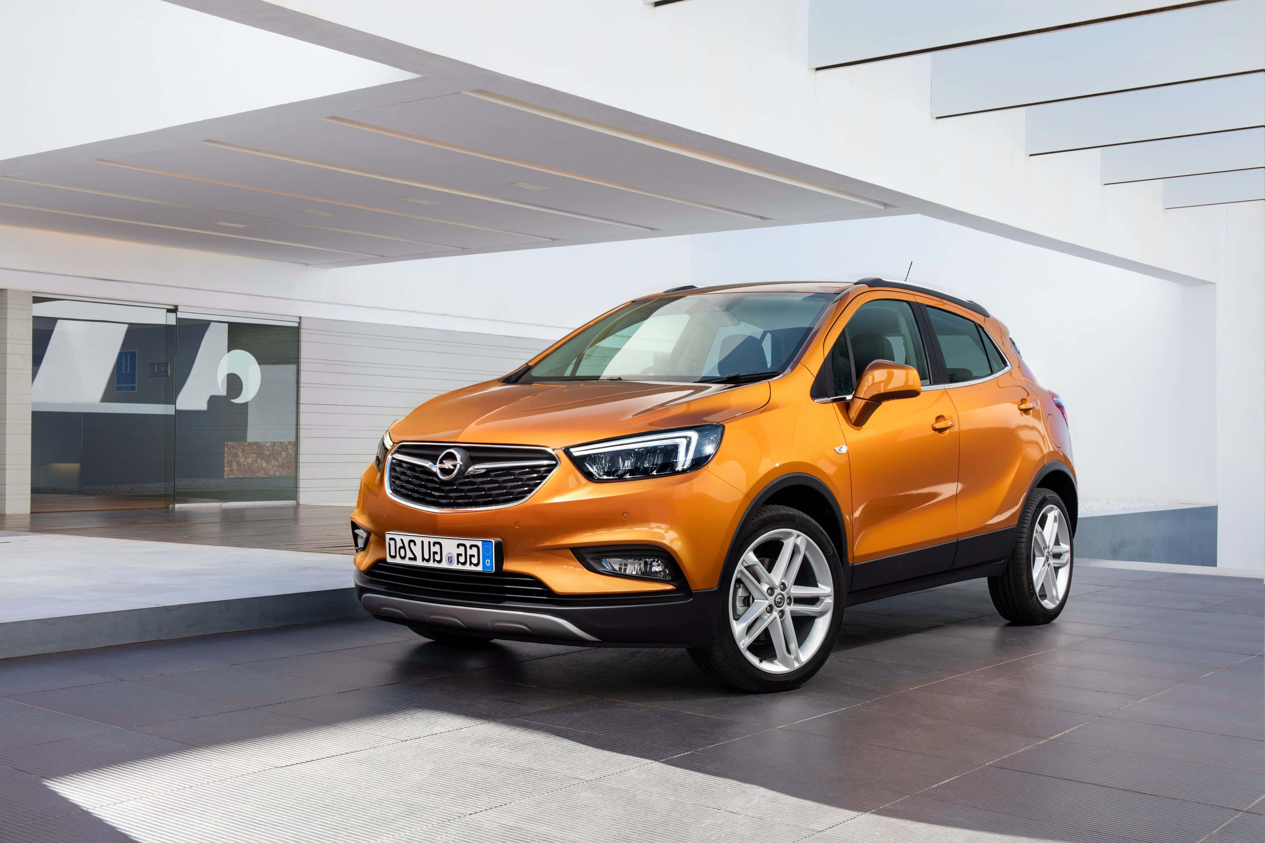 Opel Mokka 桌布 壁紙 — 一輛充滿活力的橙色 Opel Mokka SUV 停放在一個現代白色建築結構下，該結構具有流暢的線條和玻璃門。