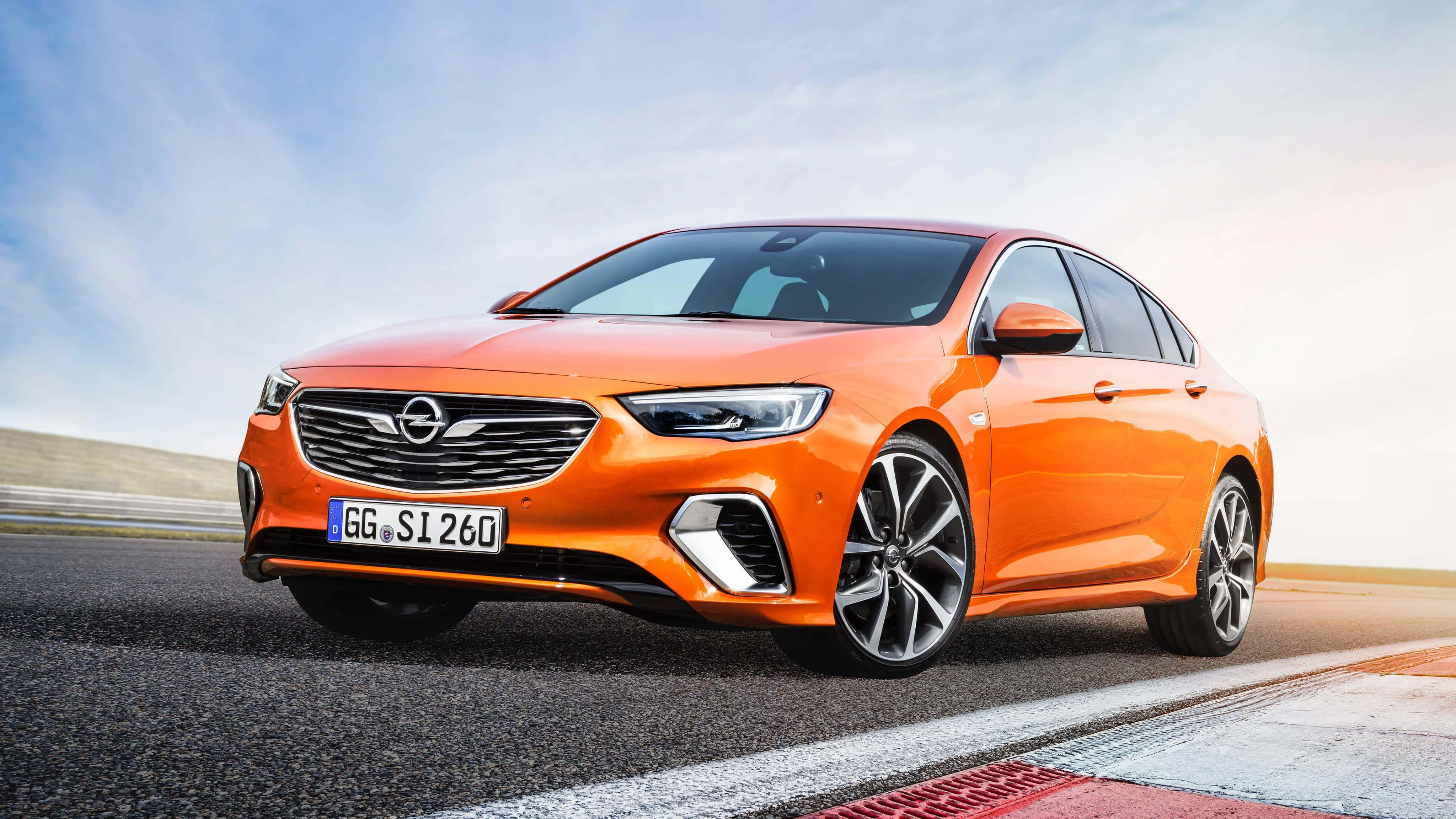 Opel Insignia 桌布 壁紙 — 一輛充滿活力的橙色歐寶轎車在晴朗的藍天白雲下沿著賽道奔馳。