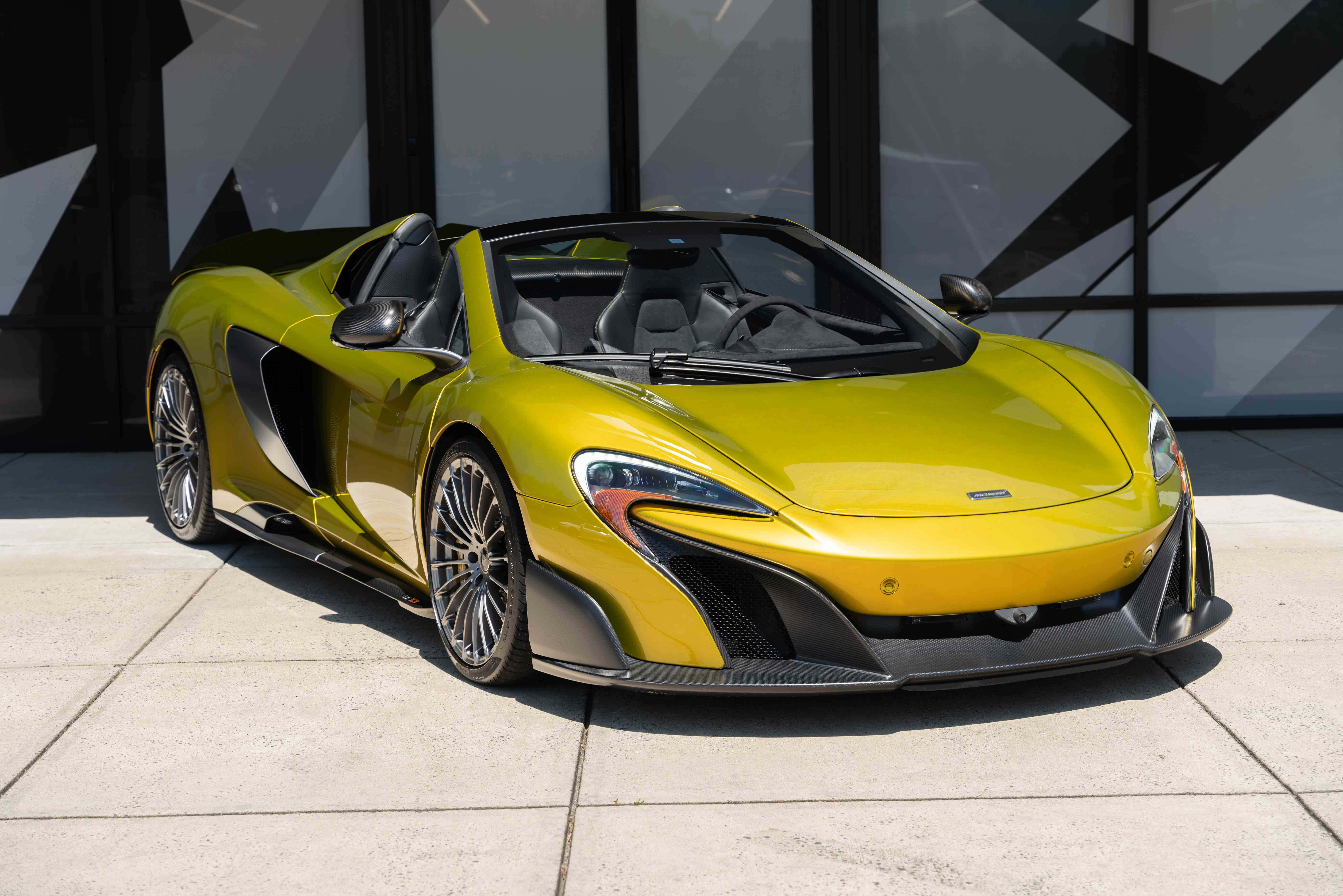 Mclaren 675lt Spider 桌布 壁紙 — 一輛充滿活力的黃色McLaren跑車，帶有黑色軟頂，停放在現代玻璃建築前的瓷磚廣場上。