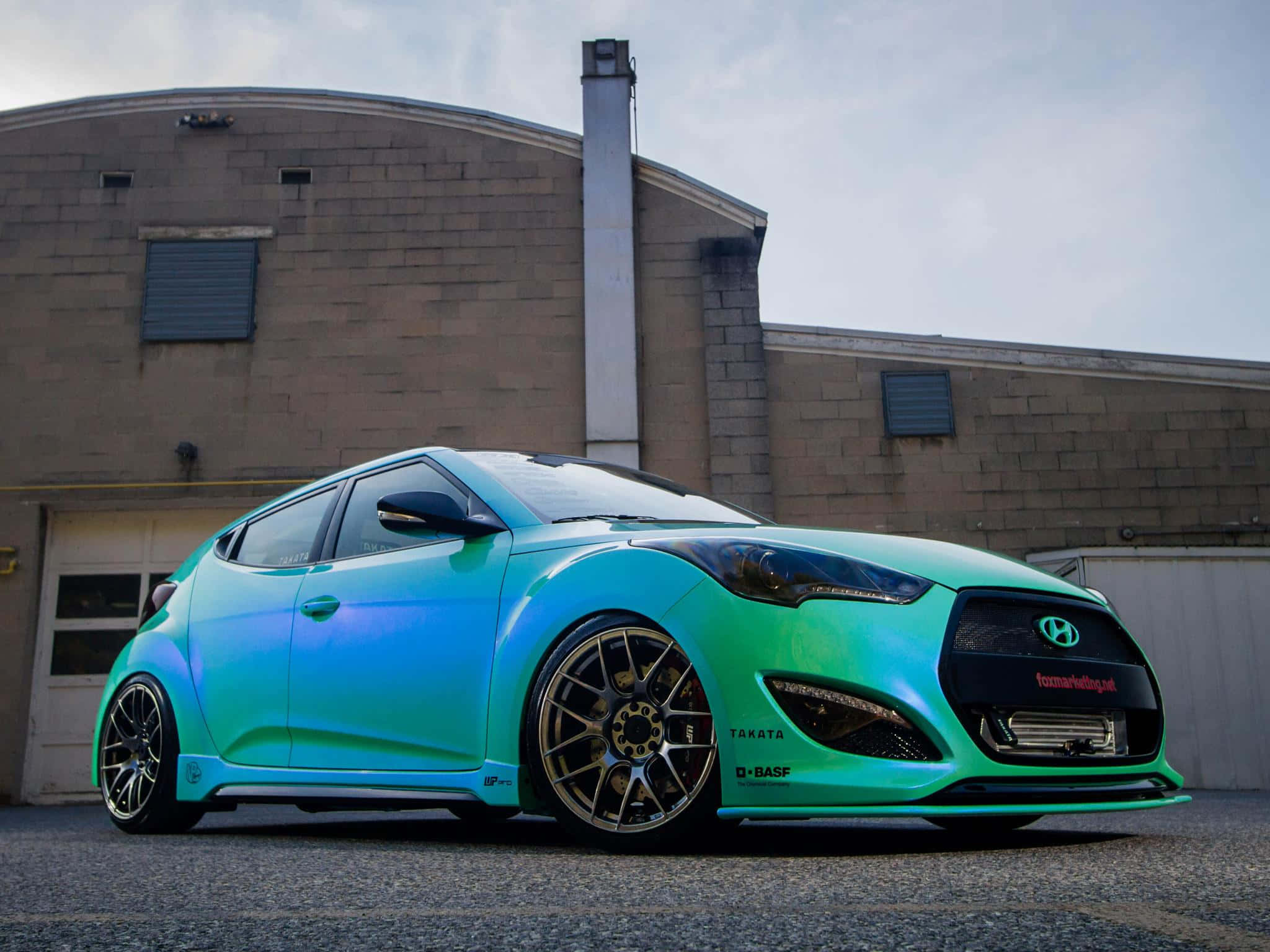 Hyundai Veloster 桌布 壁紙 — 一輛充滿活力的漸層青綠色和藍色改裝現代Veloster停在一棟磚造工業建築前。