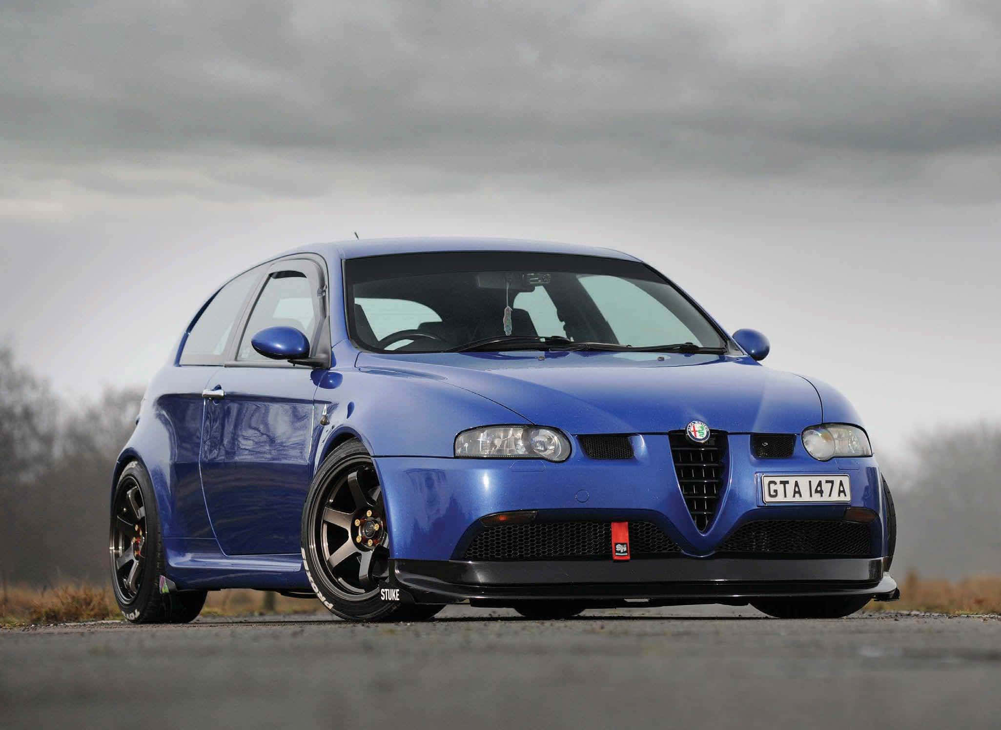 Alfa Romeo Gt 桌布 壁紙 — 一輛充滿活力的藍色愛快羅密歐GTA跑車，配備黑色輪圈，停泊在灰色瀝青路上，天空多雲。