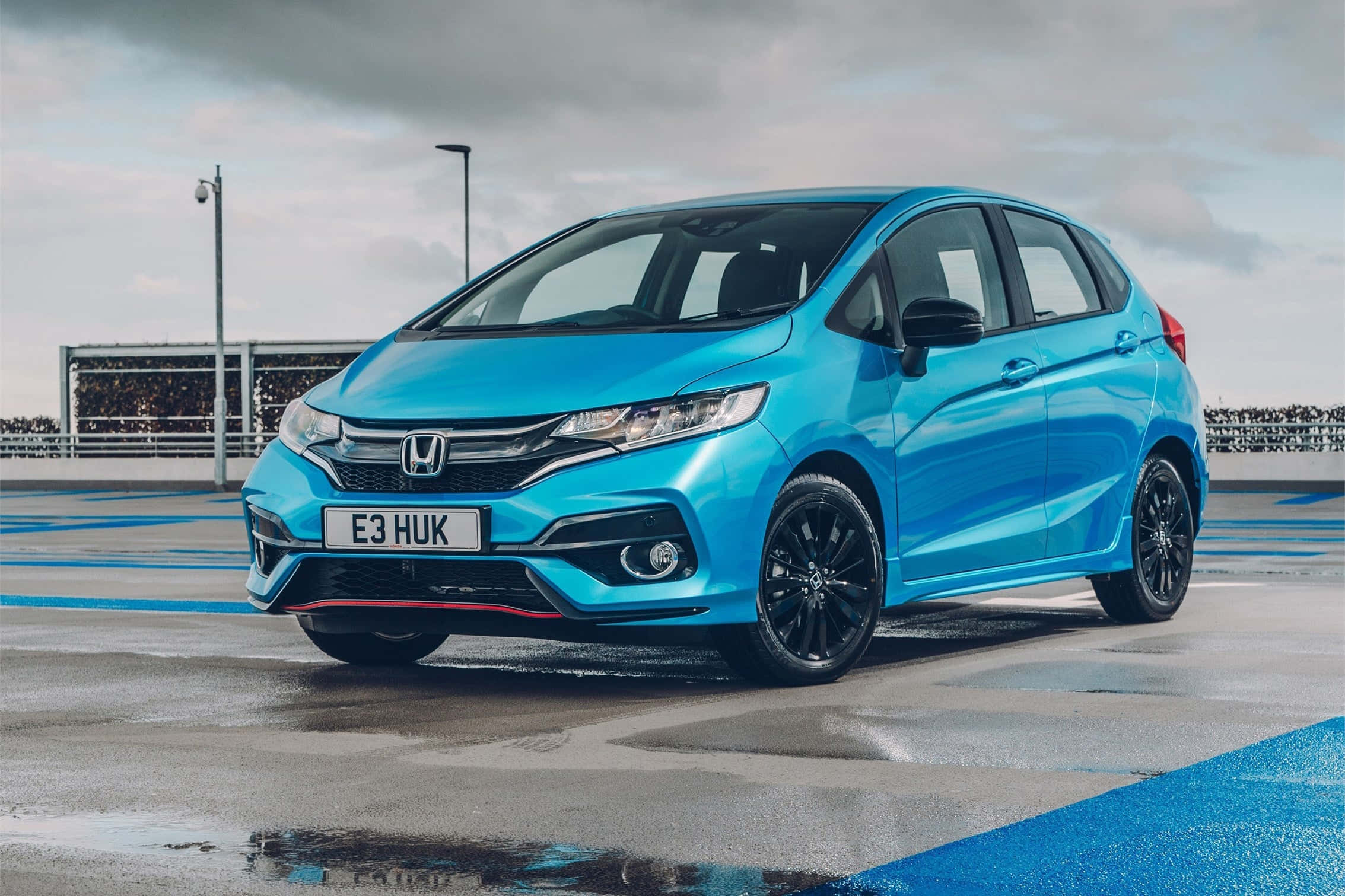 Honda Fit 桌布 壁紙 — 一輛充滿活力的藍色本田 Jazz，配黑色輪圈，停泊在潮濕的賽車場上，天空多雲。